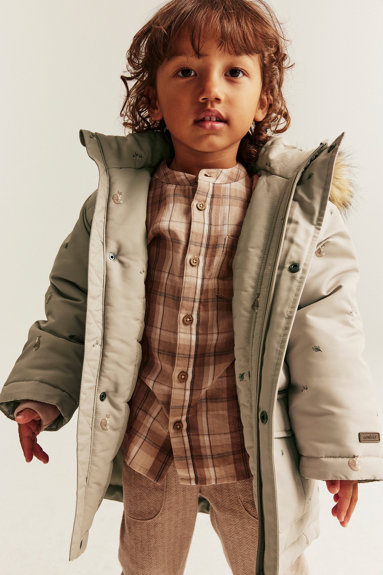 Vorderansicht eines Kindes in offener beiger Steppjacke mit Fuchsmuster über kariertem Hemd und beiger Hose.