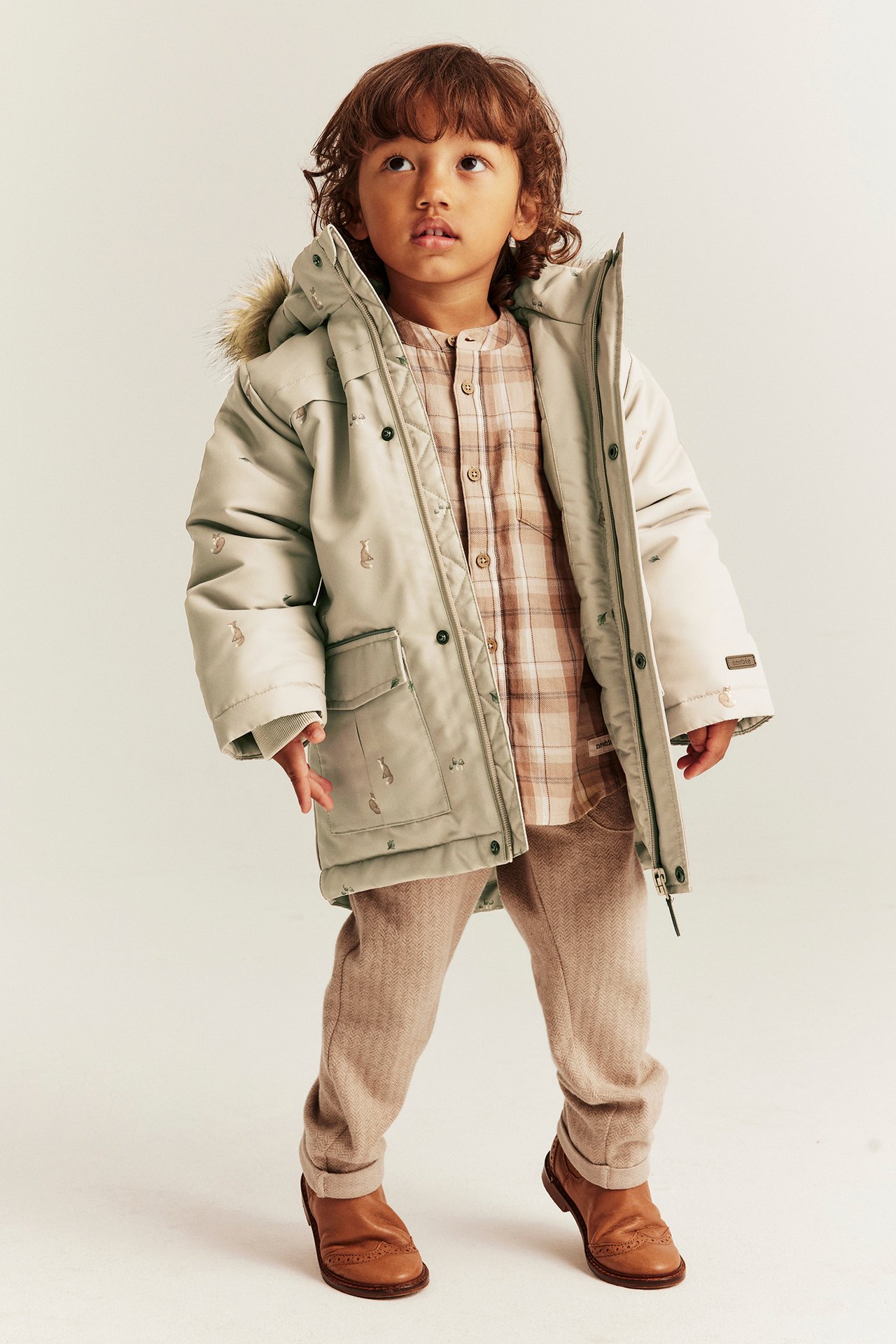 Vorderansicht: Kind in beiger, gefütterter Fuchsjacke, kariertem Hemd, brauner Hose und Brogues.
