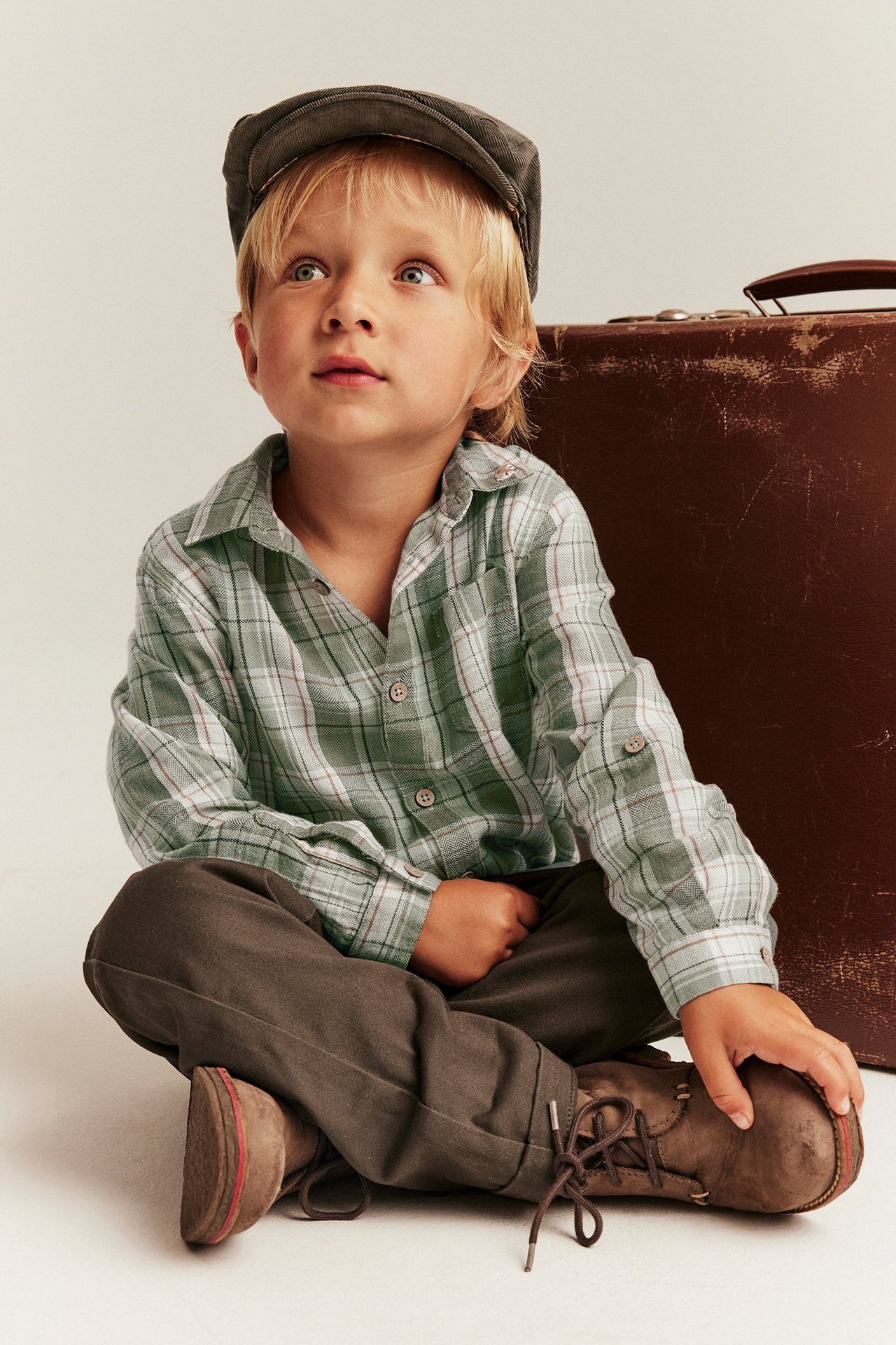 Vorderansicht: Kind trägt dunkelgrünes kariertes Baumwollhemd, dunkelbraune Hose, Mütze und braune Schuhe.
