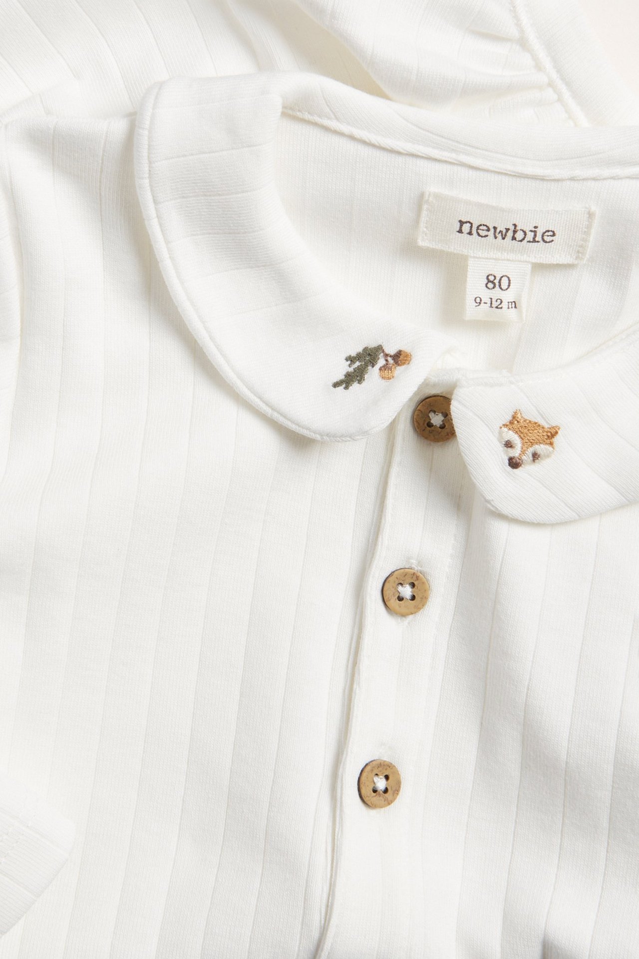 Nahaufnahme eines gerippten, cremefarbenen Baby-Bodys mit langen Ärmeln, Kragen und Fuchs- sowie Eichelstickerei.