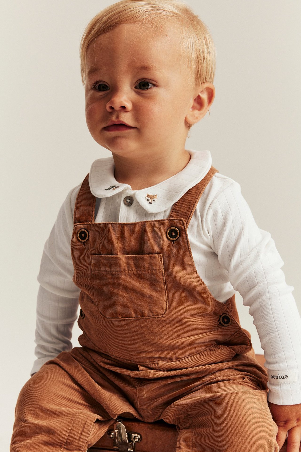 Close-up front view of a baby wearing an off-white ribbed bodysuit with fox embroidery and brown corduroy overalls.