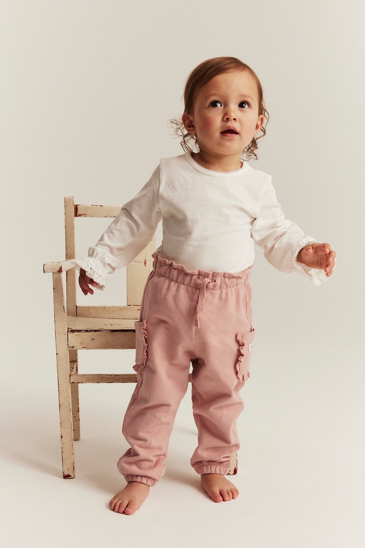 Vorderansicht eines Babys in rosa Baumwoll-Jogginghose mit Rüschen-Taschen und weißem Langarmshirt.
