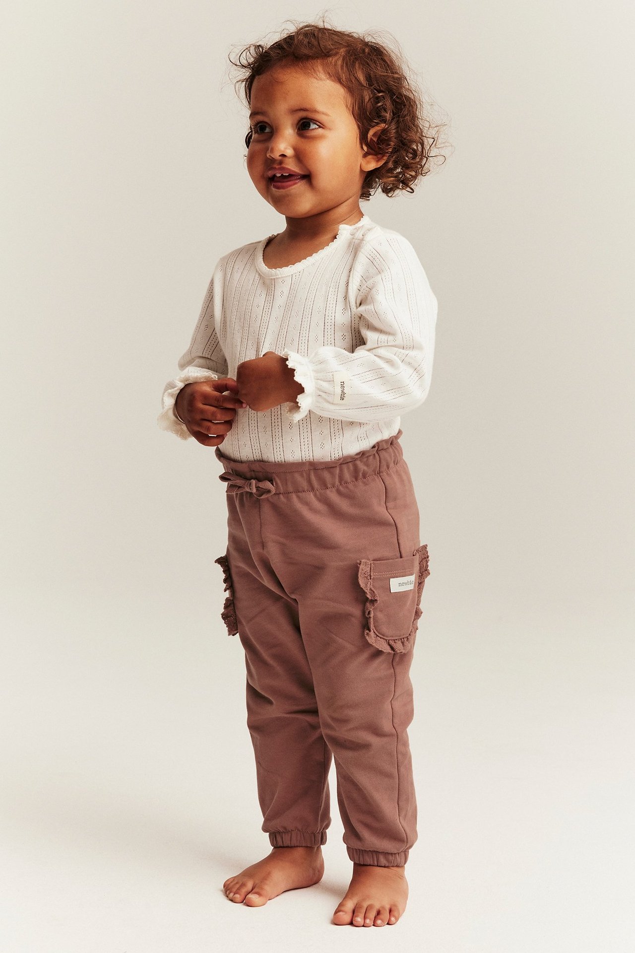 Vorderansicht: Baby in dunkelrosa Baumwoll-Jogginghose mit Rüschentaschen und weißem Langarmshirt.
