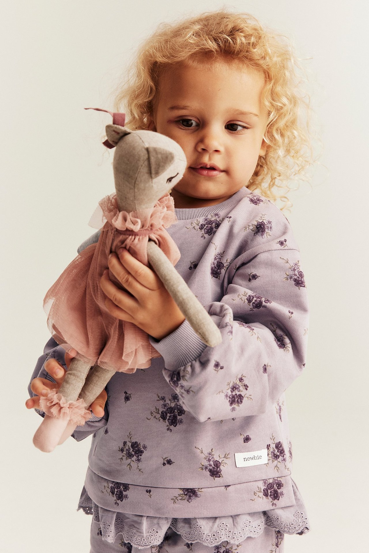 Vorderansicht eines Kindes in helllila geblümtem Baumwoll-Sweatshirt mit Spitzenrüsche, hält eine Stoffpuppe.