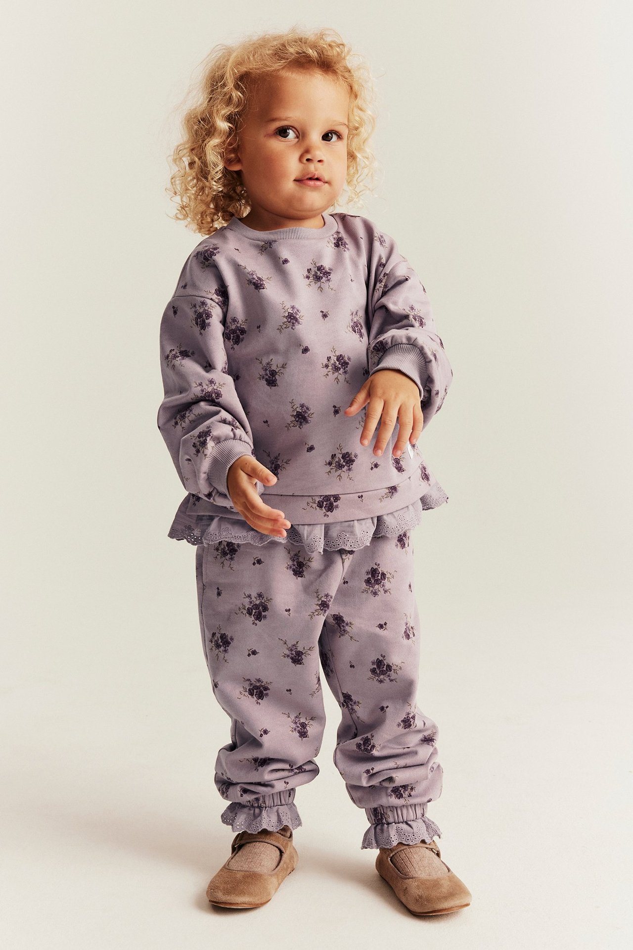 Vorderansicht eines Kindes in helllila Jogginghose mit Blumenmuster, Spitzenrüschen und passendem Sweatshirt.