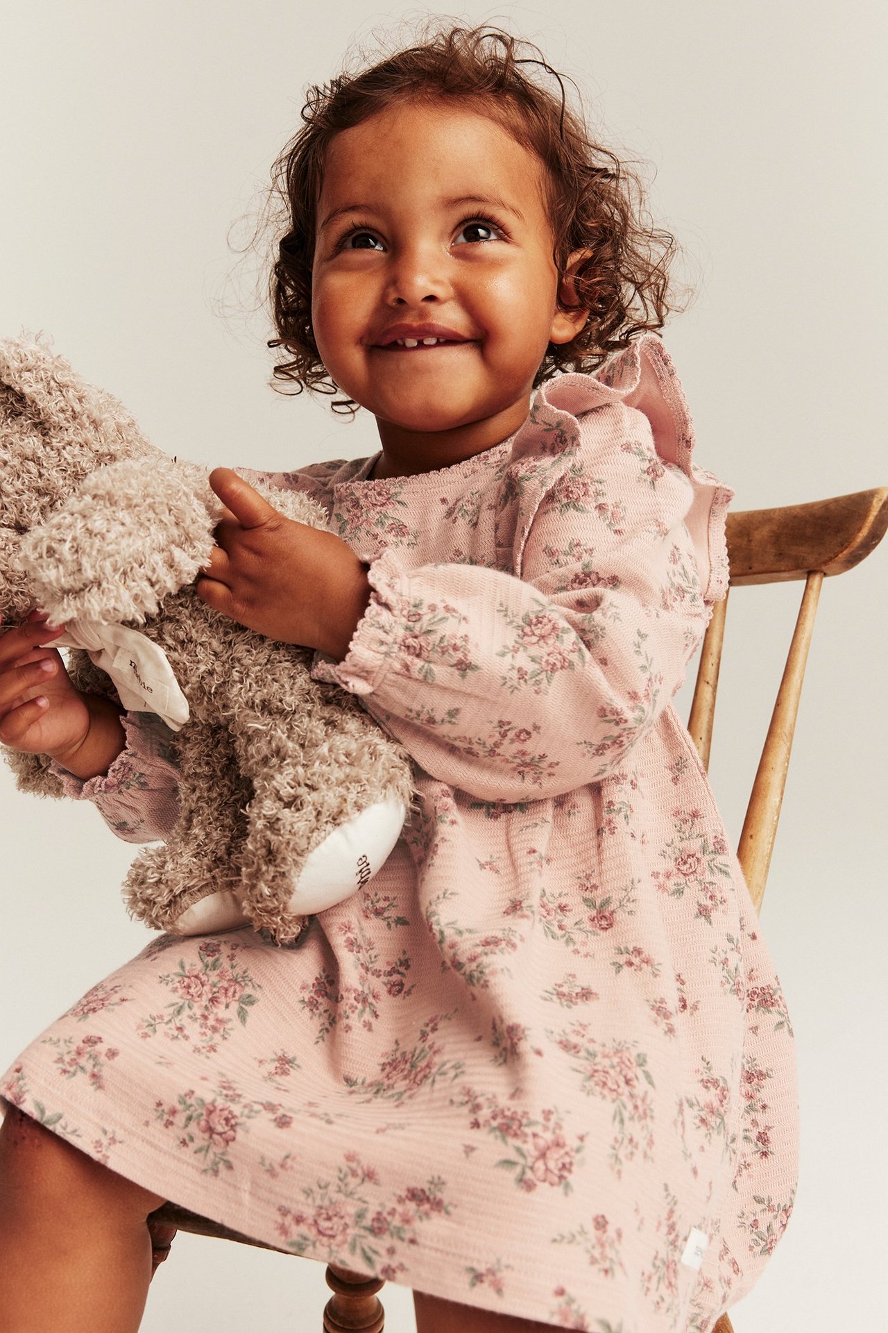 Vorderansicht eines Kindes in rosa geblümtem Jerseykleid mit Rüschen, hält ein braunes Stofftier.
