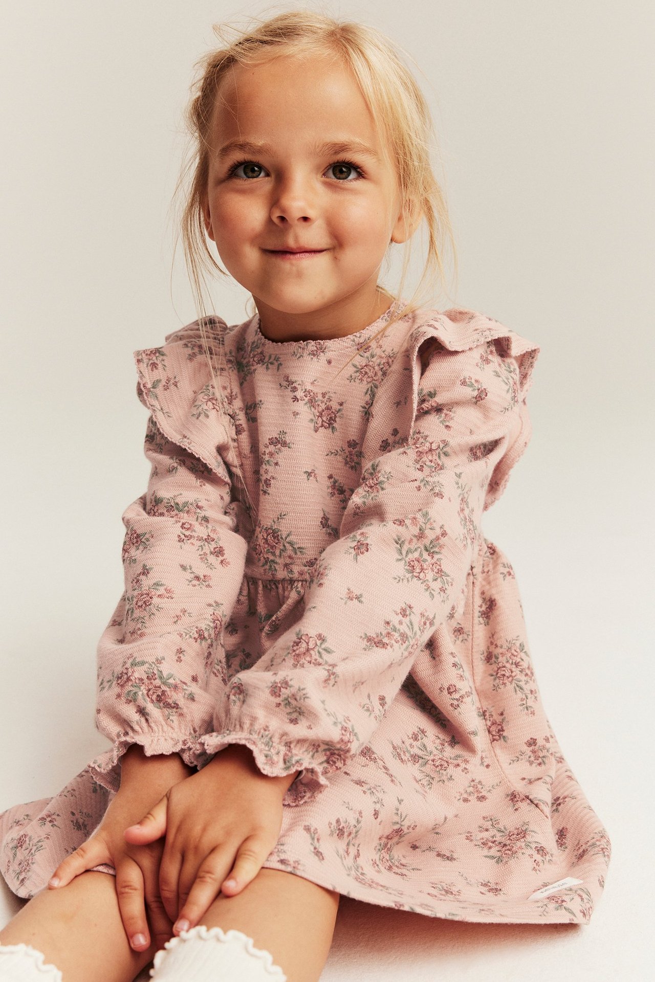 Front view of a kid wearing a pink floral structured jersey dress with ruffles and white ruffled socks.