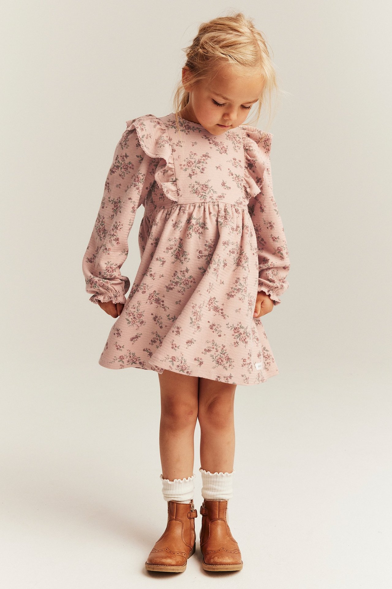 Front view of a kid wearing a pink floral dress with ruffles, white socks, and brown boots.