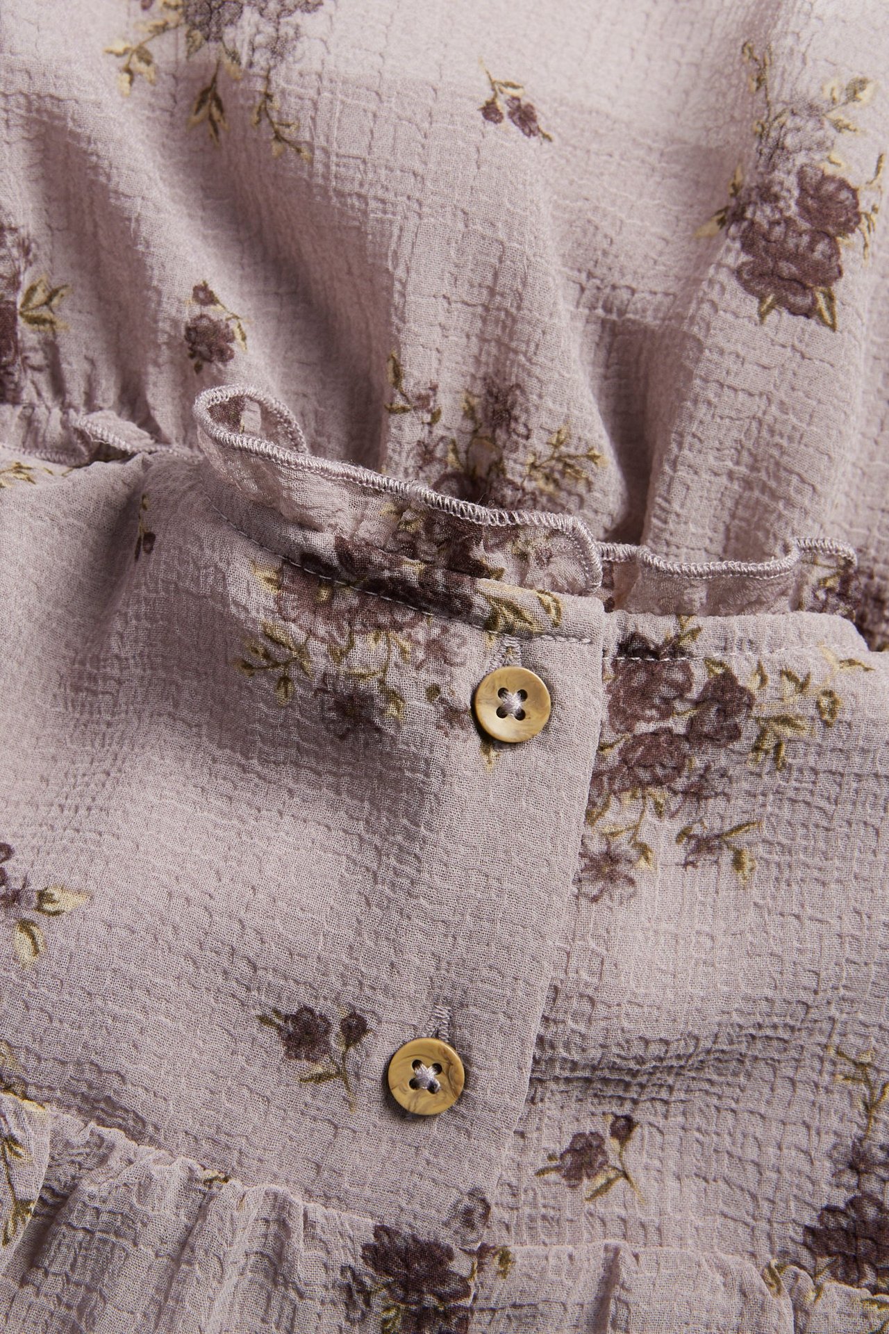 Nahaufnahme eines helllila geblümten Chiffonkleides mit Rüschen und zwei hellbraunen Knöpfen.