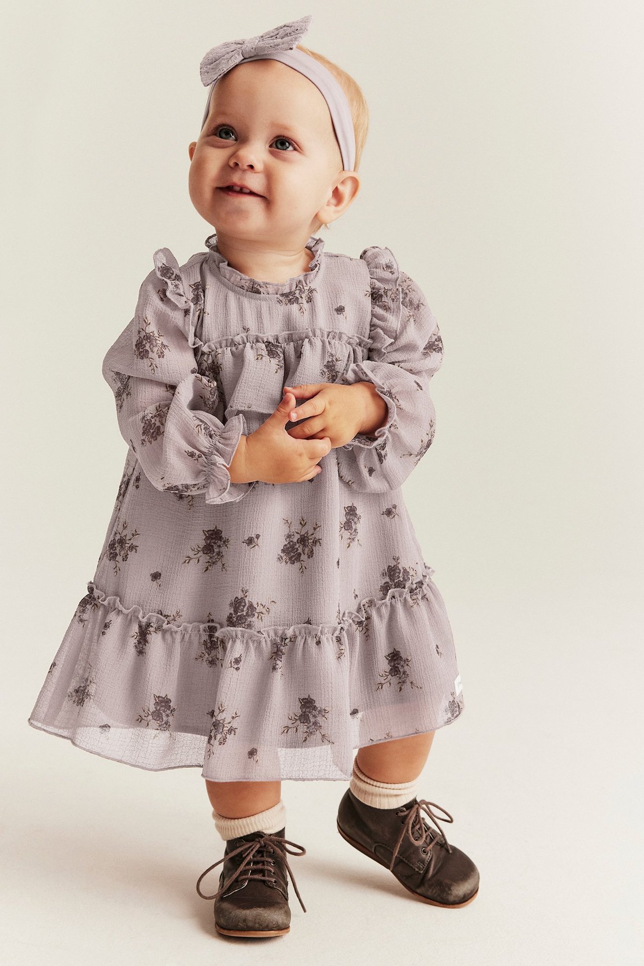 Vorderansicht: Baby in helllila geblümtem Chiffonkleid mit Rüschen, passendem Haarband, weißen Socken, braunen Schuhen.