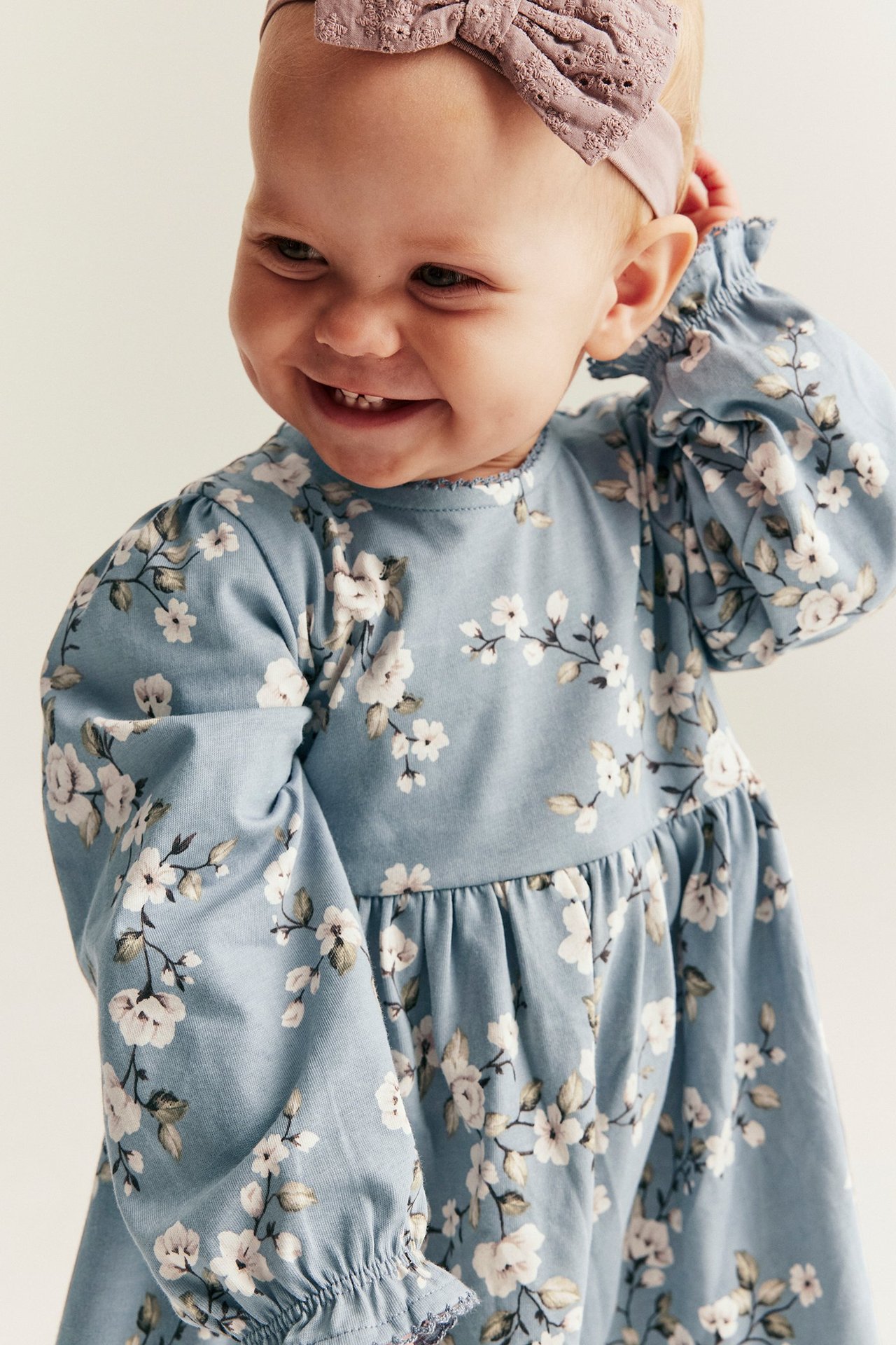 Nahaufnahme eines lächelnden Babys in hellblauem Blumen-Jerseykleid mit Puffärmeln und lila Spitzenstirnband.