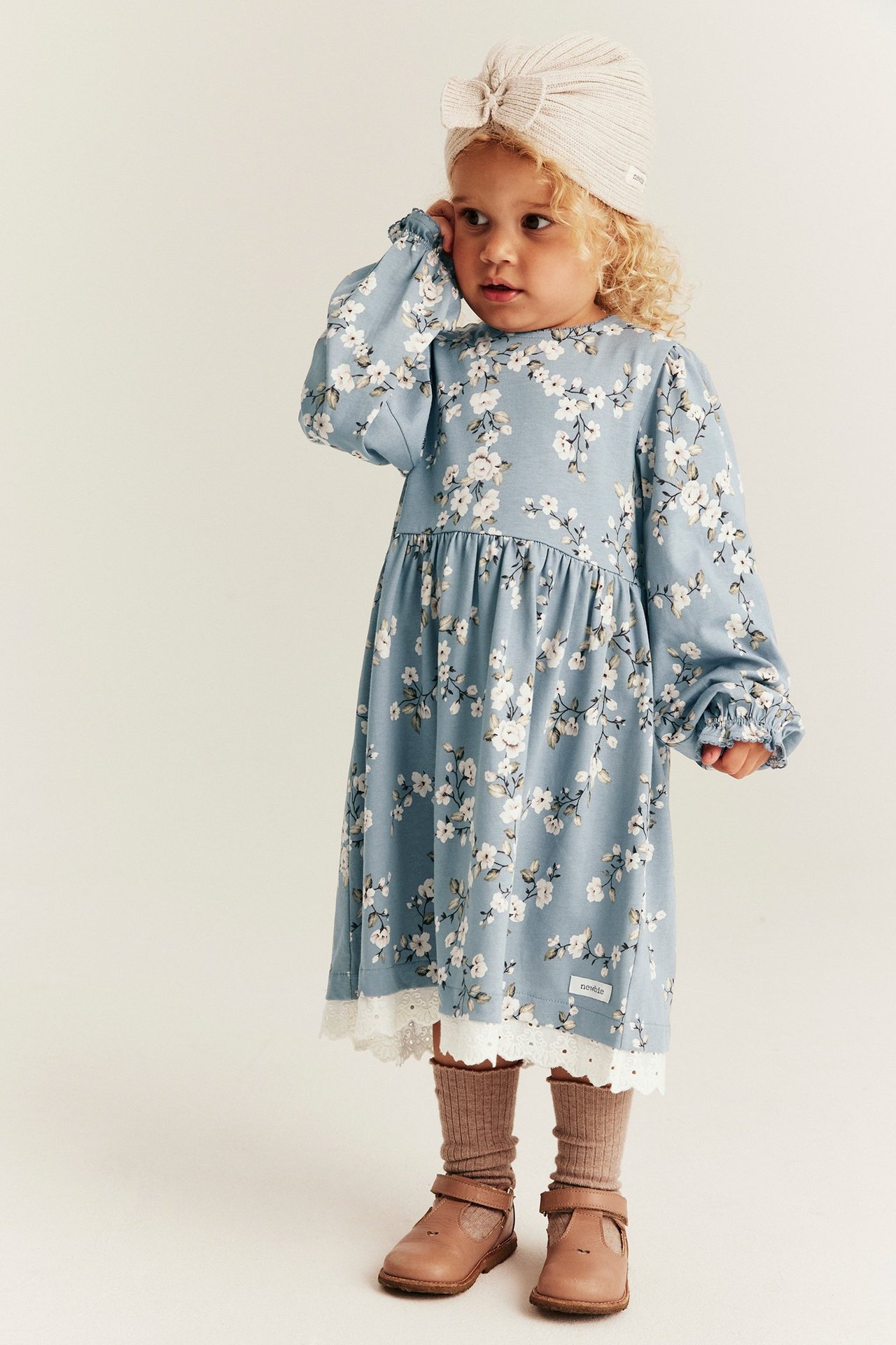 Front view: Kid in a light blue floral jersey dress with lace, beige turban, brown ribbed socks, and brown shoes.