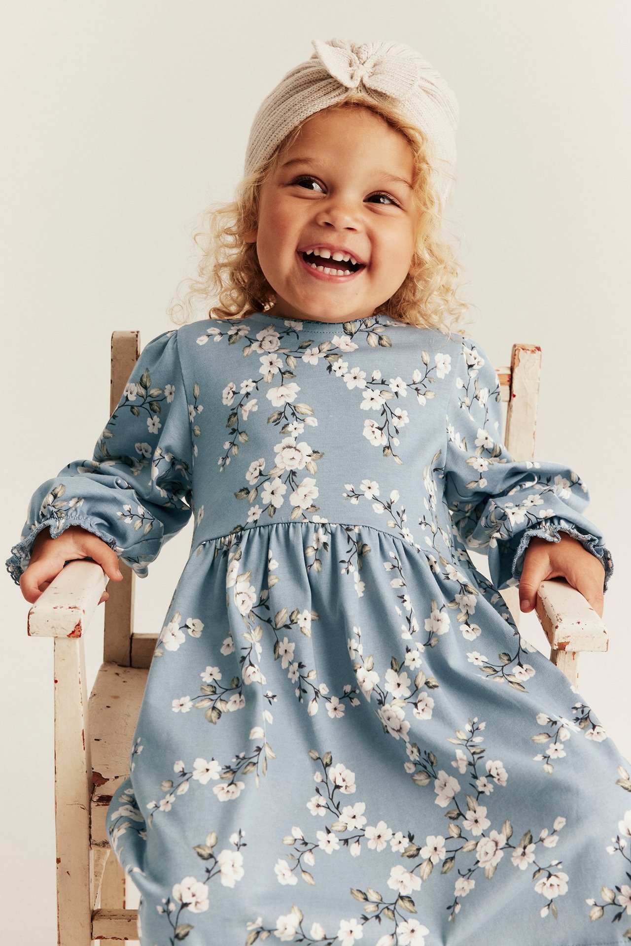 Front view of a smiling kid wearing a light blue floral jersey dress and a light beige ribbed headband.
