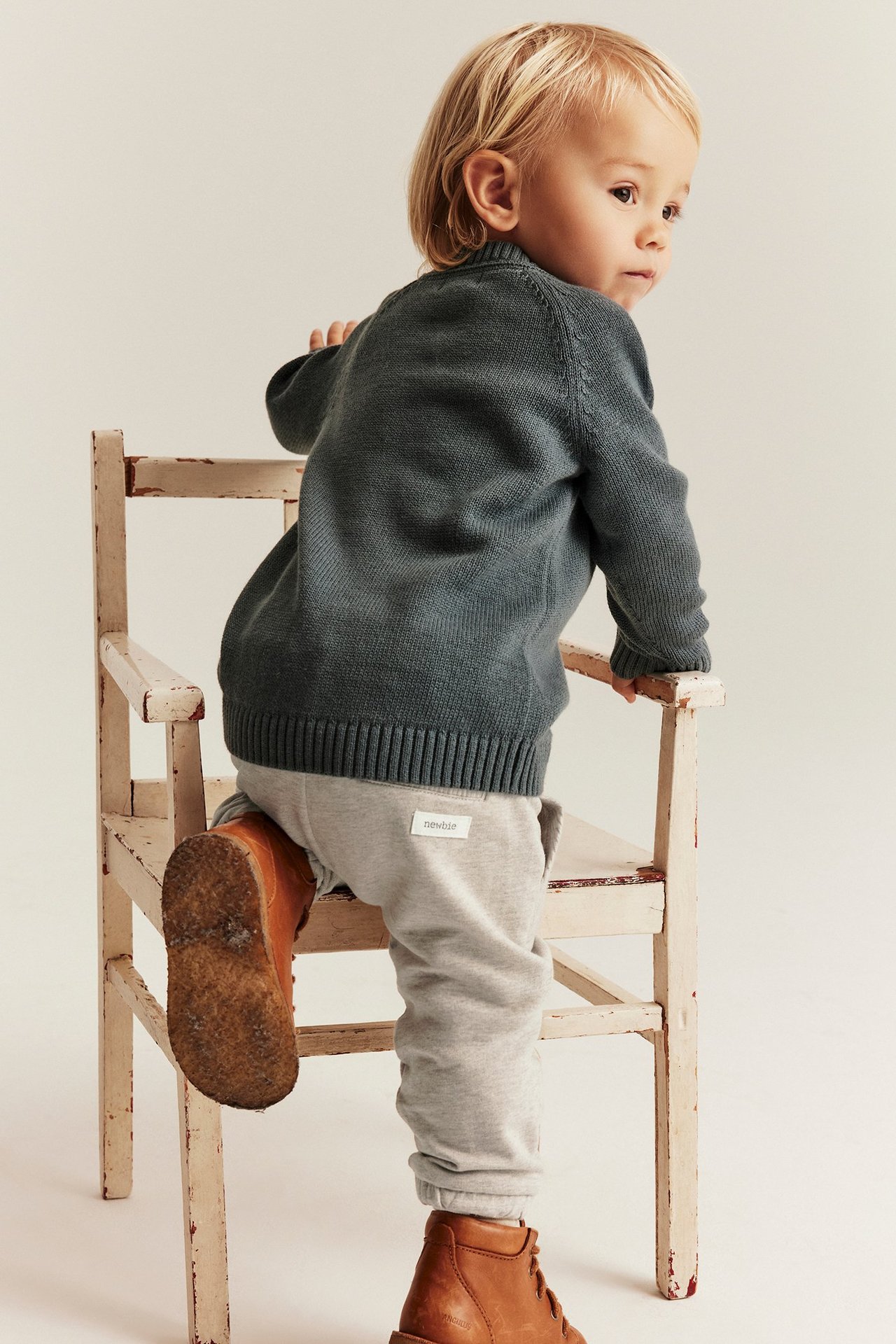 Back-side view: baby in light beige melange joggers, dark grey knitted sweater, and brown boots, standing on chair.