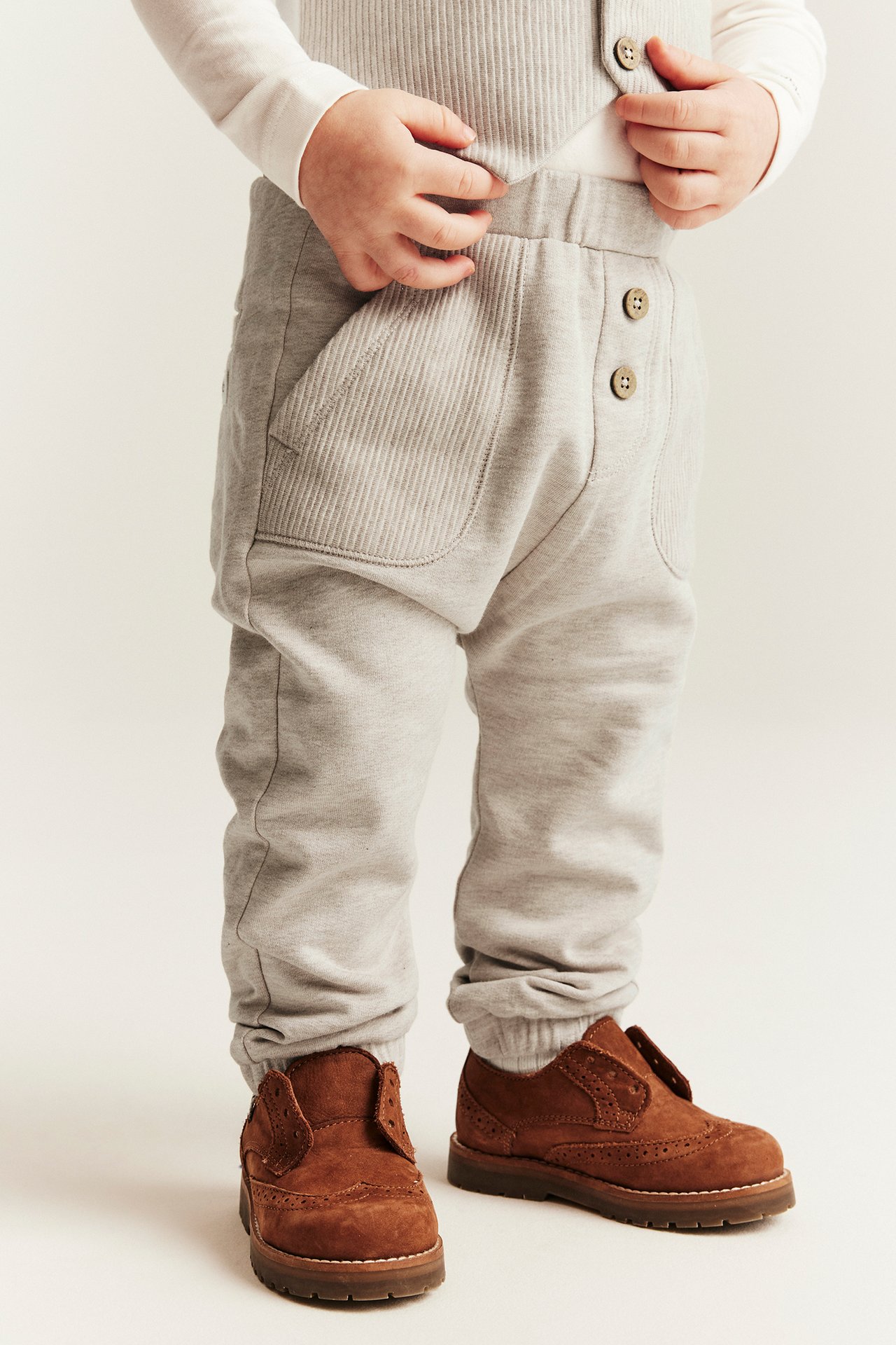 Front view: baby in beige melange cotton joggers with pockets, buttons, white top, and brown shoes.