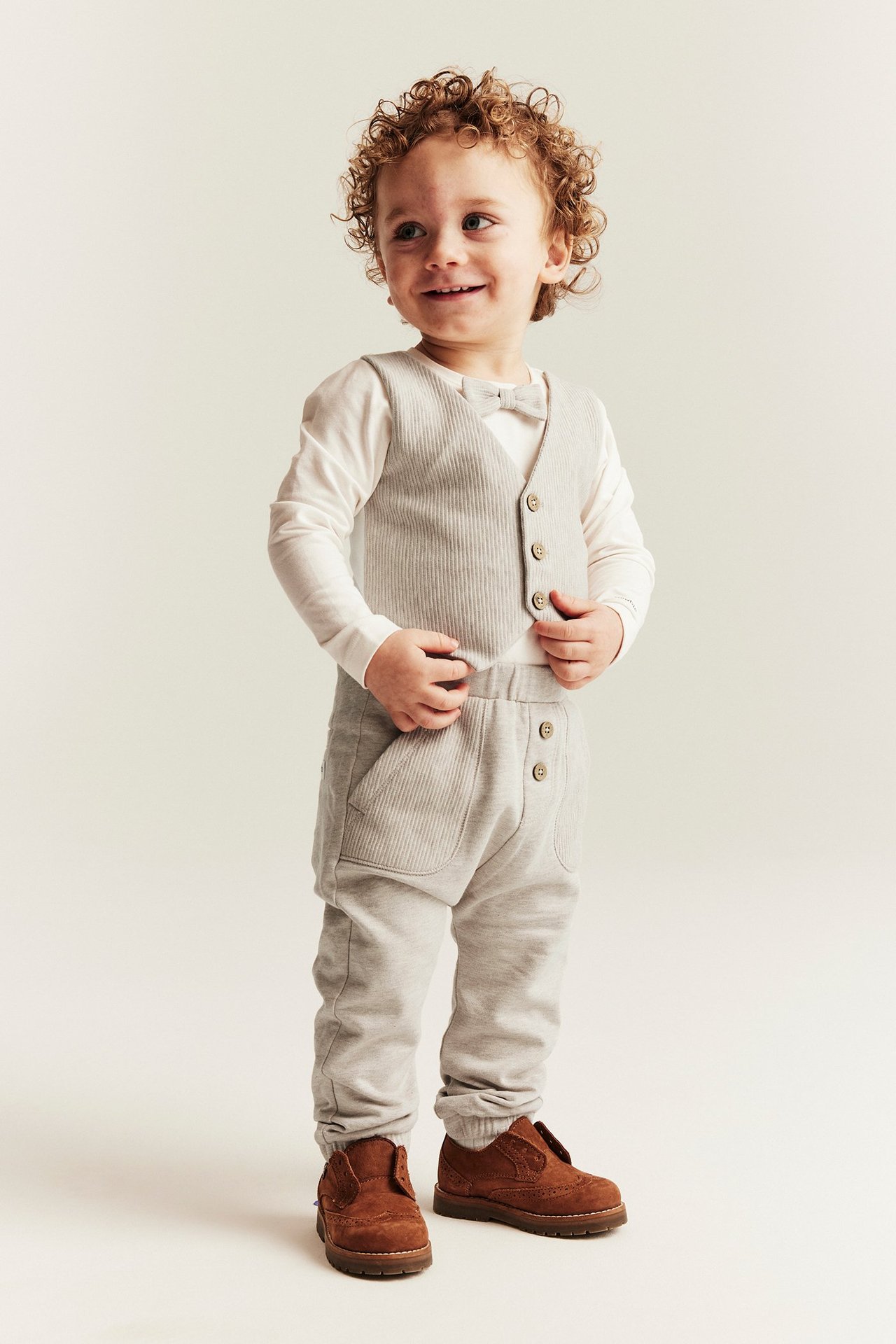 Full-body: baby boy in beige melange cotton sweatpants, ribbed vest, white top, and brown shoes.
