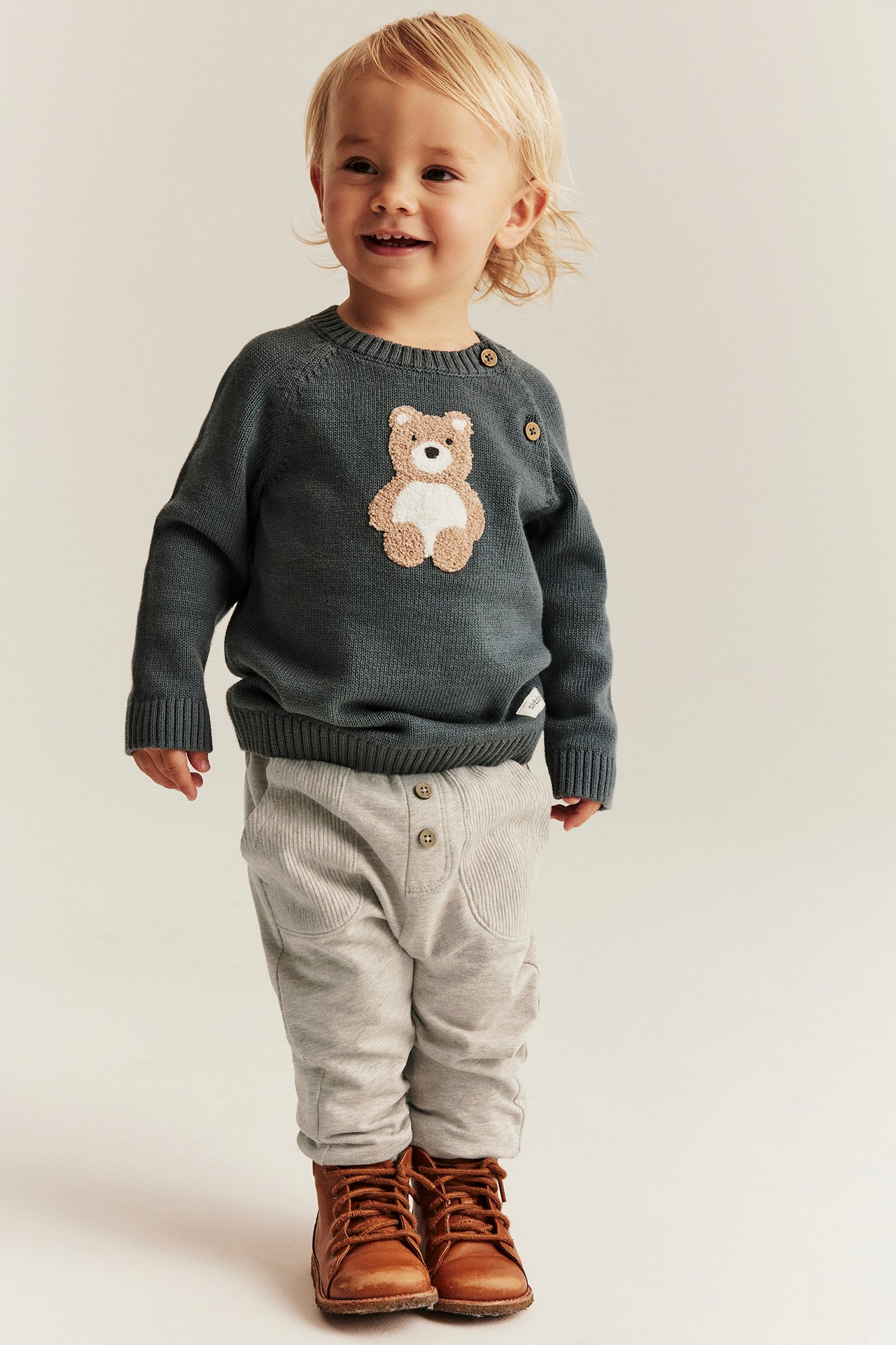 Baby in beige melange jogging pants, dark sweater with teddy bear, and brown boots.