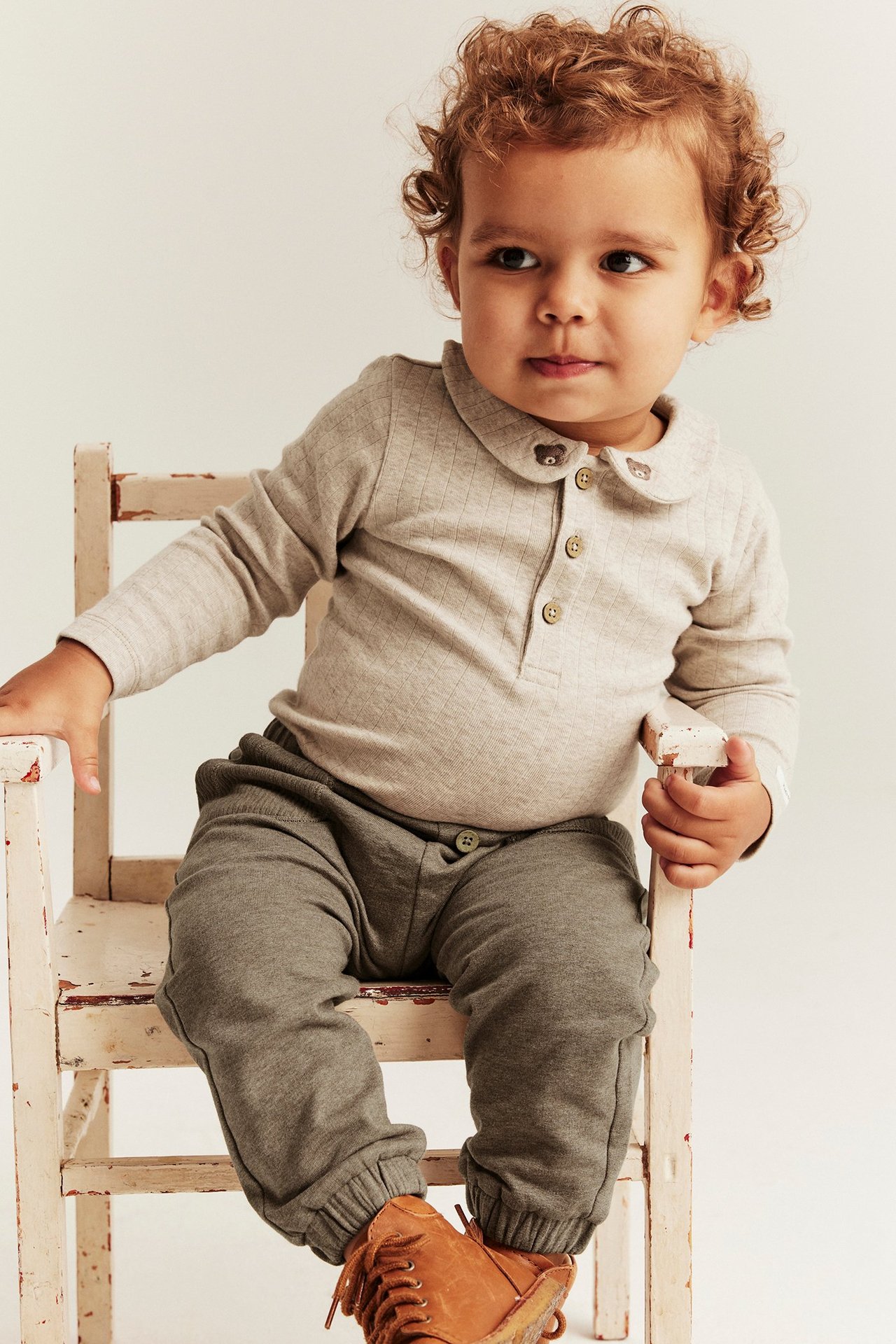 Front view: Baby in brown melange jogging pants with buttons, a light grey ribbed top with bear collar, and brown shoes.
