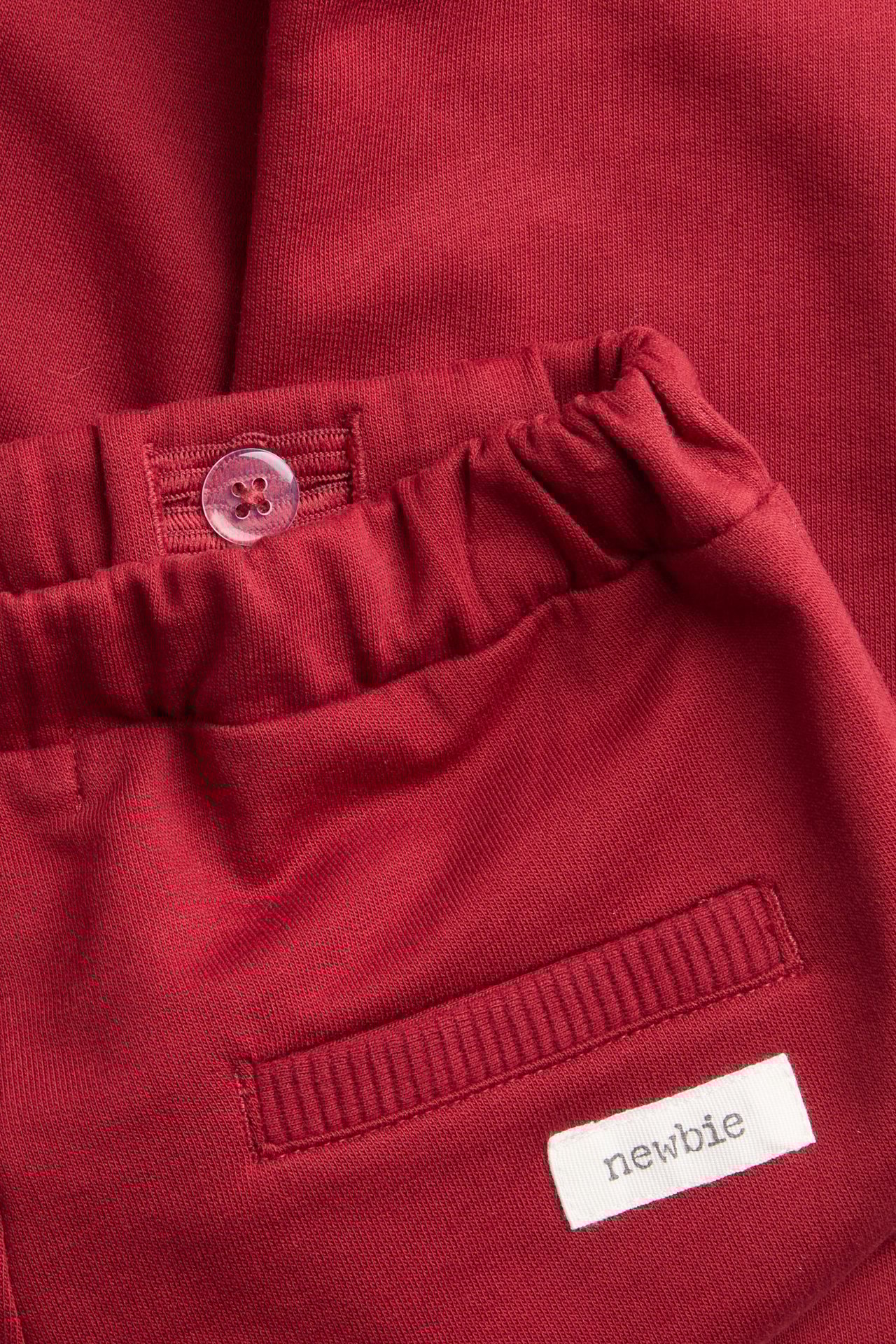 Nahaufnahme roter Baumwoll-Jogginghose mit elastischem Bund, Zierknopf, gerippter Fake-Tasche und Newbie-Etikett.