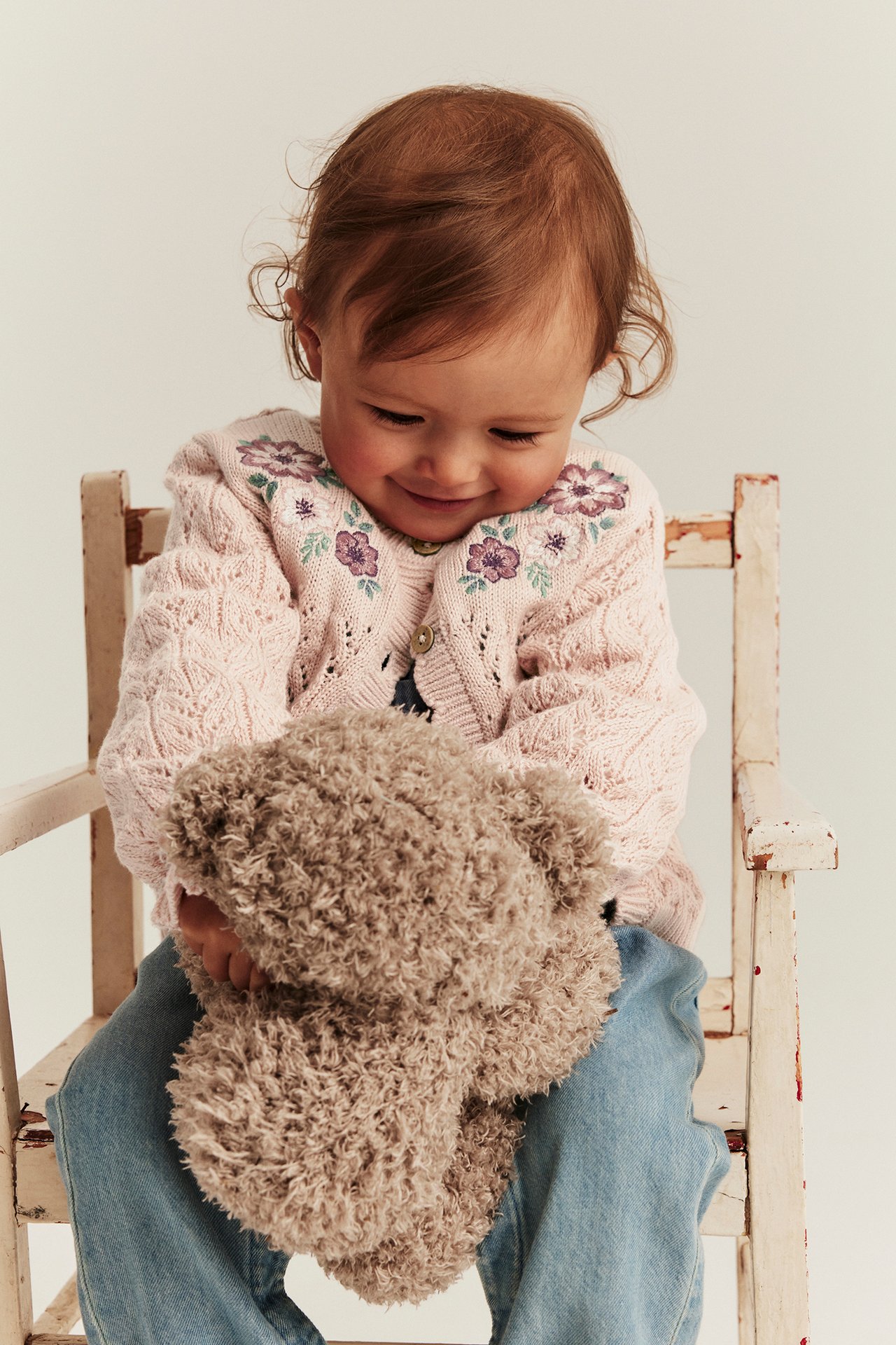 Front view: baby in a light pink embroidered knit cardigan, blue jeans, holding a brown teddy bear.