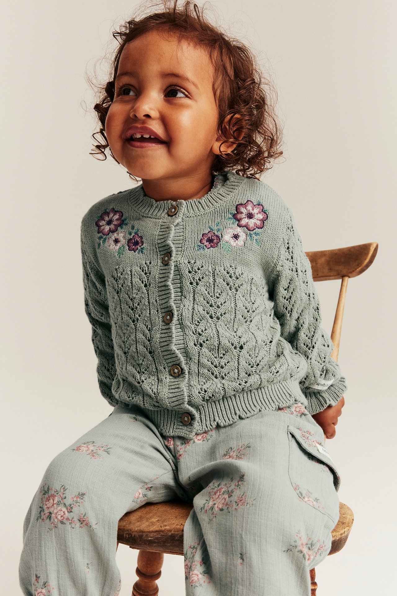 Vorderansicht eines Baby-Mädchens in hellgrüner Strickjacke mit Blumenstickerei und geblümter Hose.