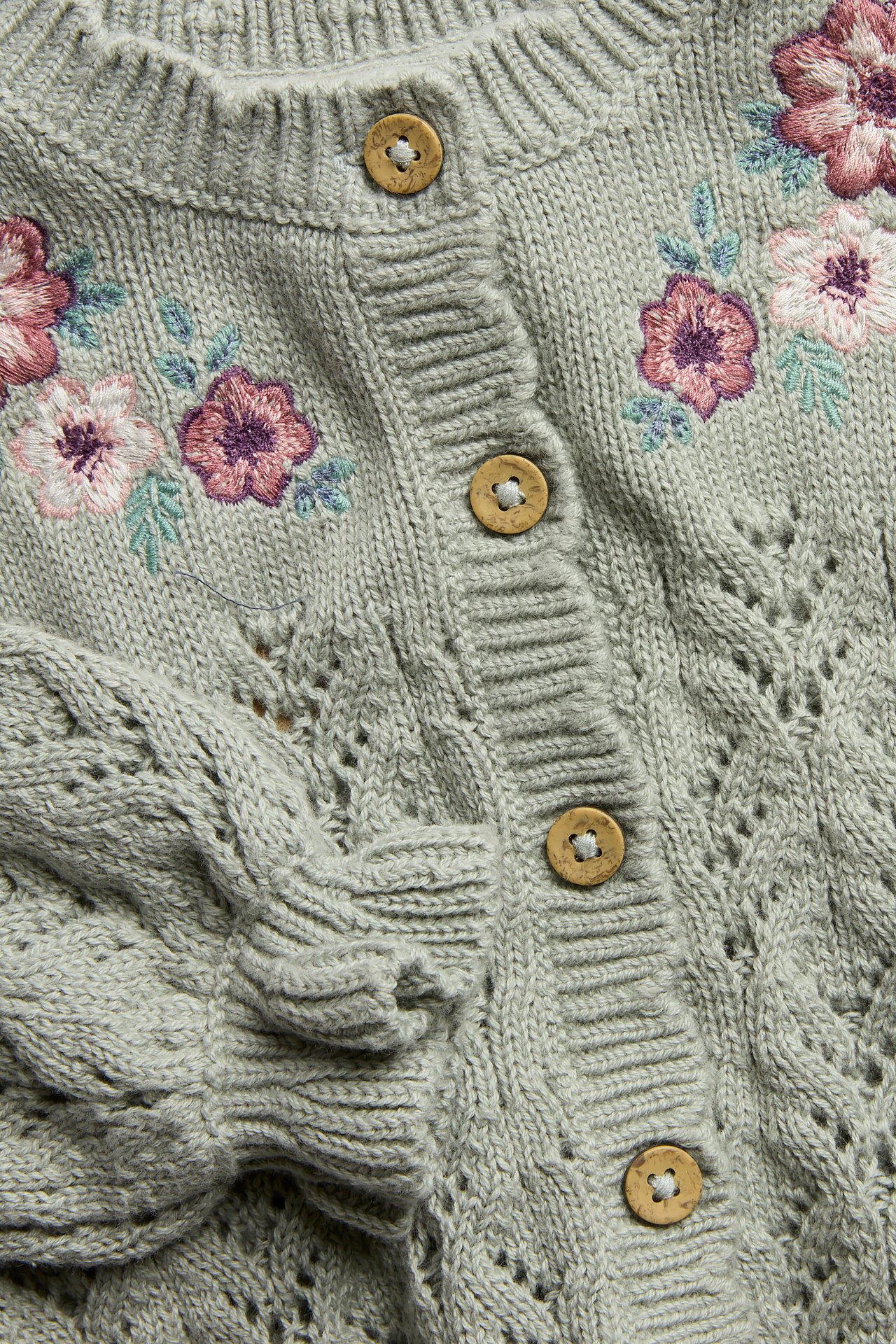 Nahaufnahme vorne einer grünen gestrickten Baumwollstrickjacke mit Blumenstickerei und Holzknöpfen.
