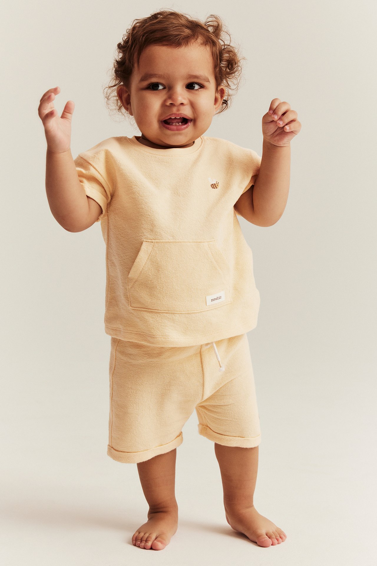 Vorderansicht: Baby in gelben Baumwollshorts mit Kordelzug und passendem T-Shirt mit Bienenstickerei.