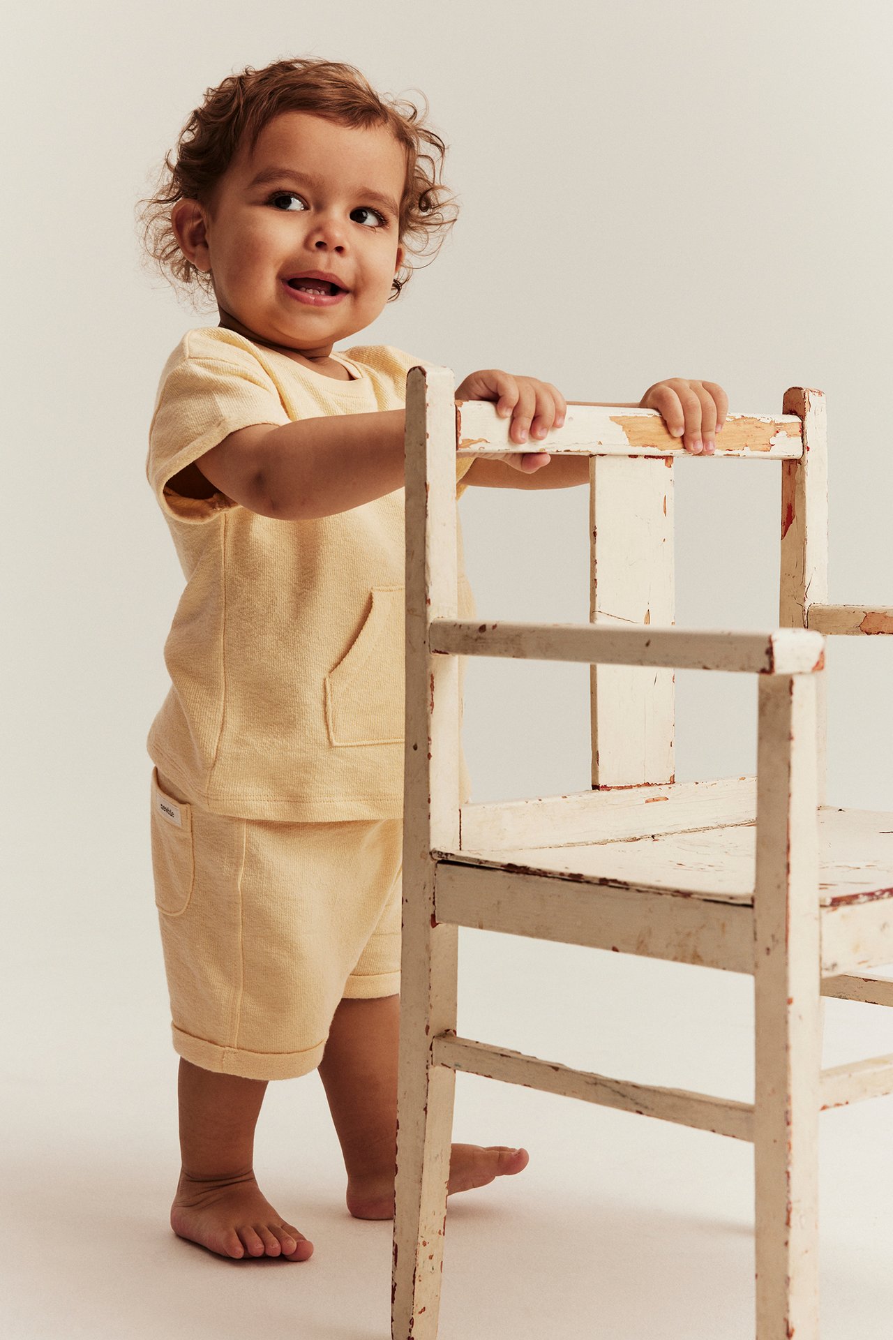 Front view: baby in yellow cotton t-shirt with pocket and matching shorts, holding distressed chair.