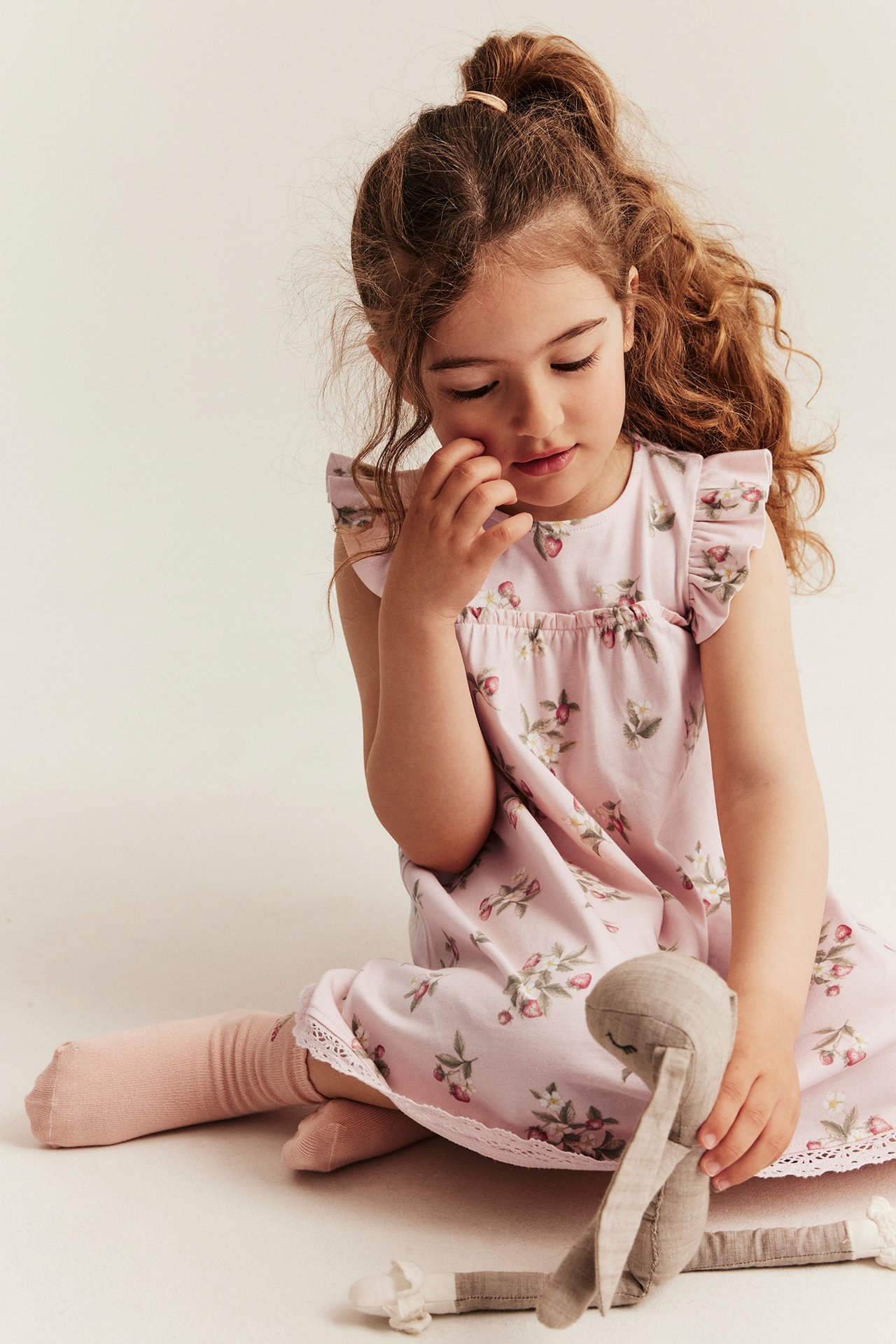 Front view: Kid in light pink patterned jersey dress, ruffle sleeves, lace hem, pink socks, holding doll.