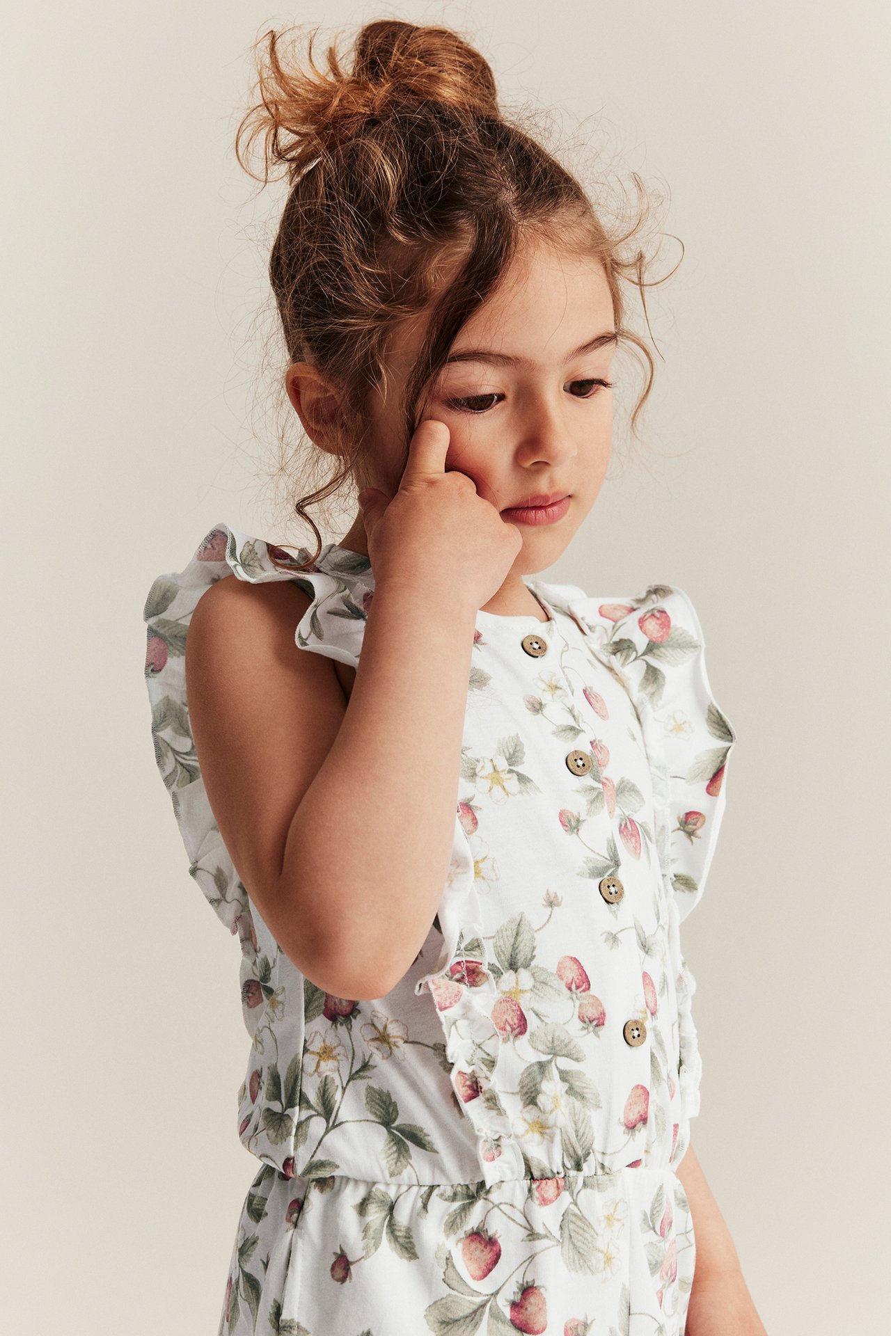 Front view of a kid wearing an off-white cotton sleeveless jumpsuit with strawberry pattern, ruffles, and front buttons.