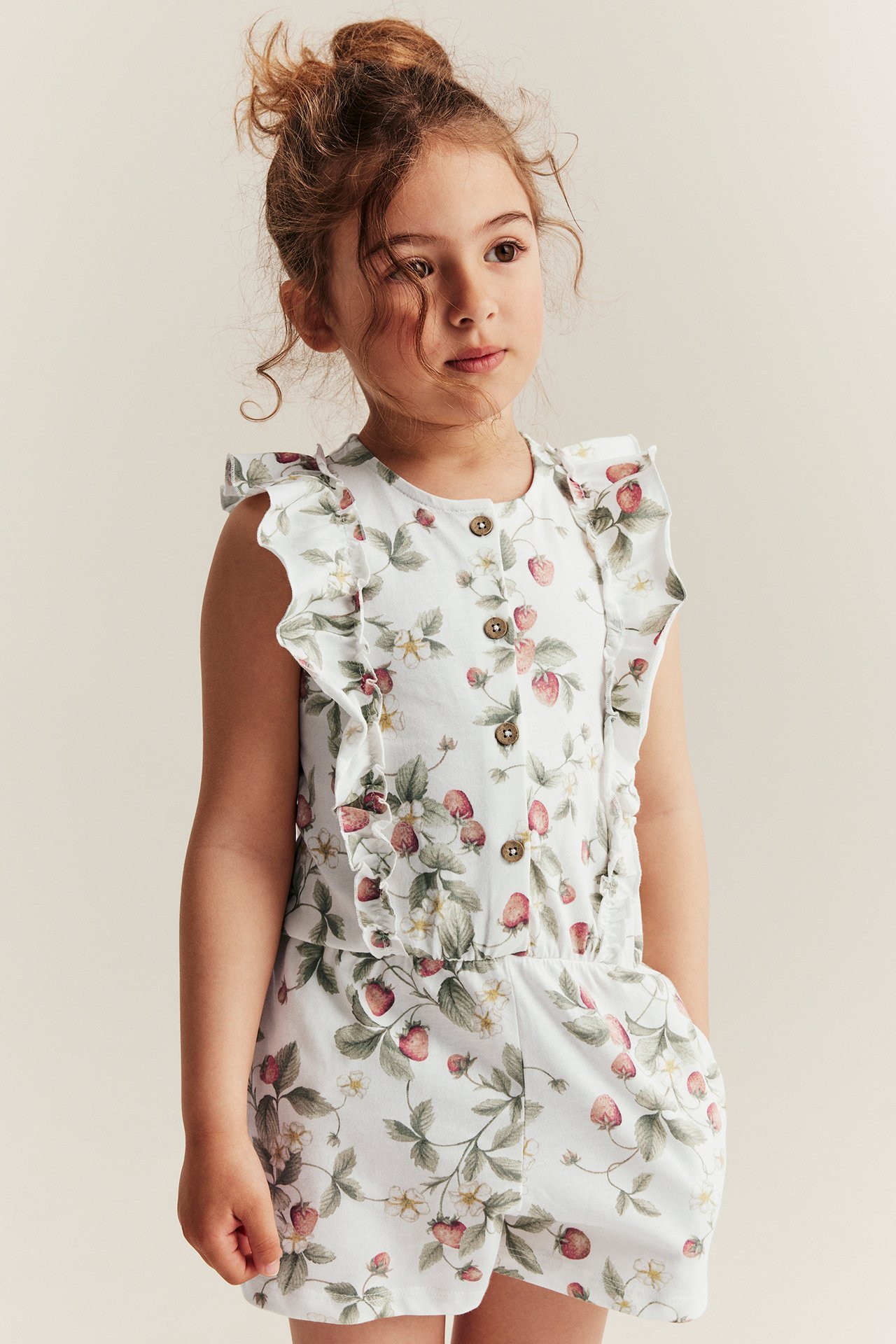 Front view: Kid in offwhite cotton jersey sleeveless jumpsuit with strawberry pattern, ruffles, short legs.