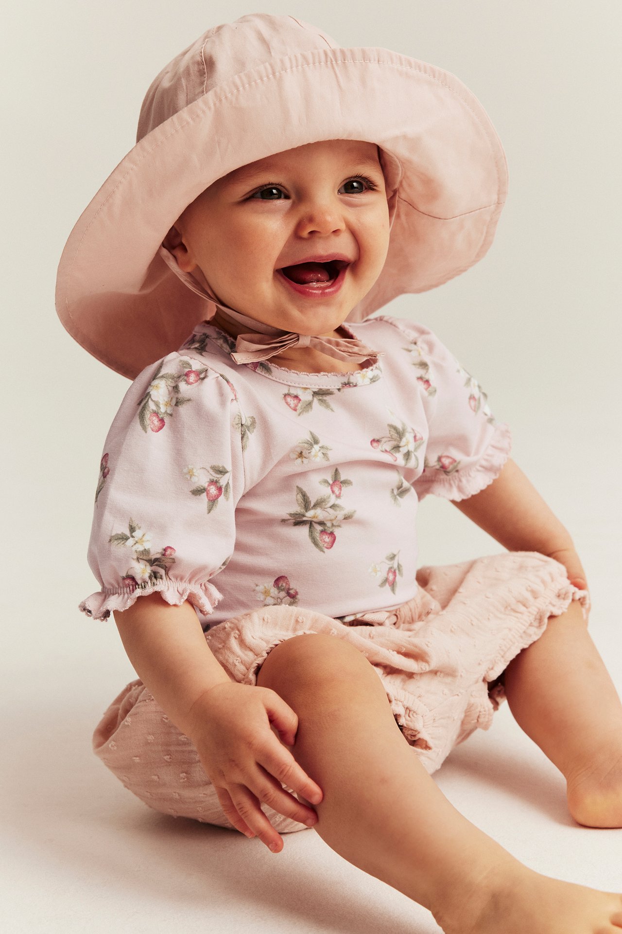 Front view of a smiling baby in a light pink strawberry-patterned bodysuit, pink bloomers, and a sun hat.