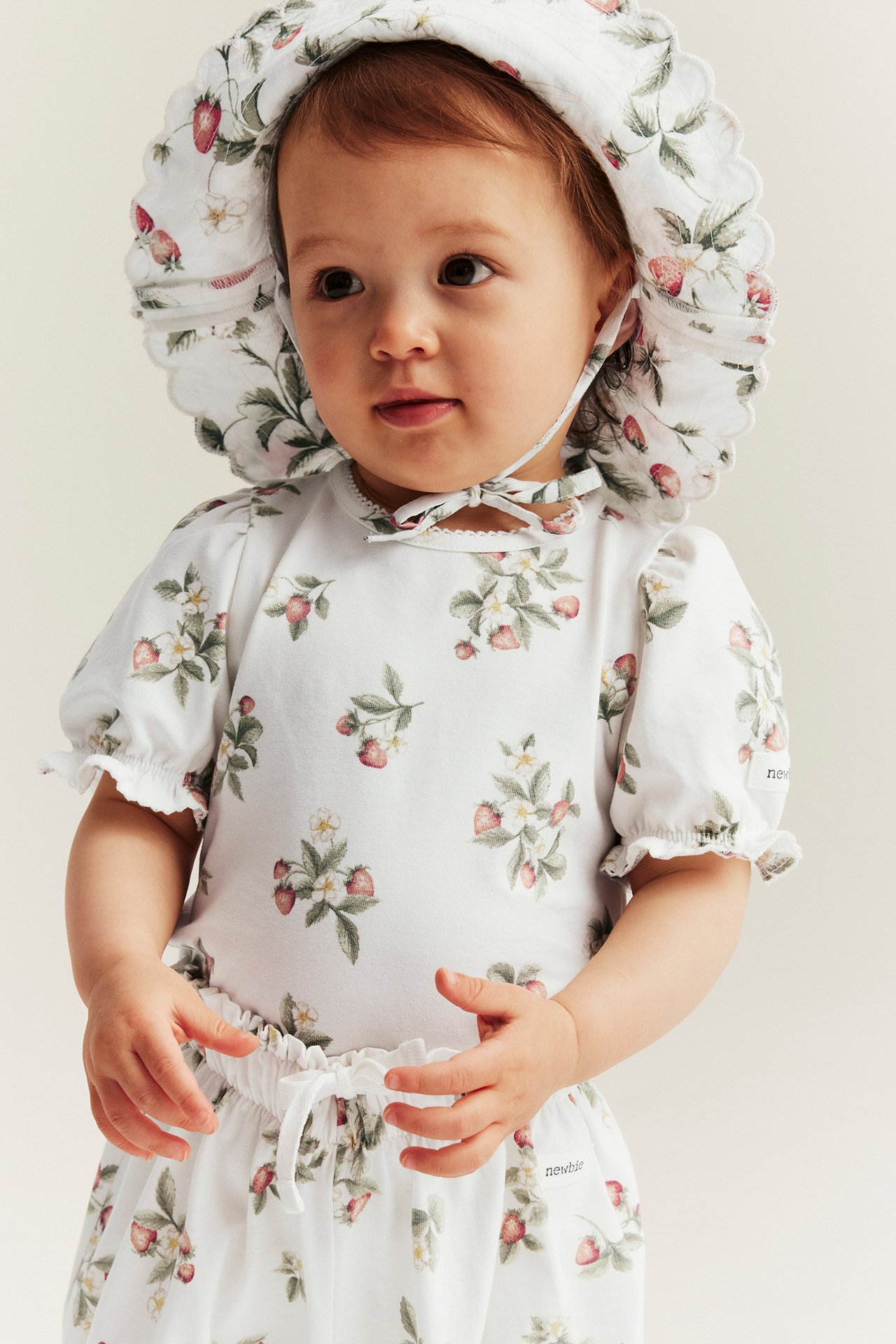 Vorderansicht eines Babys in einem cremefarbenen kurzärmeligen Body und passendem Hut mit Erdbeermuster.
