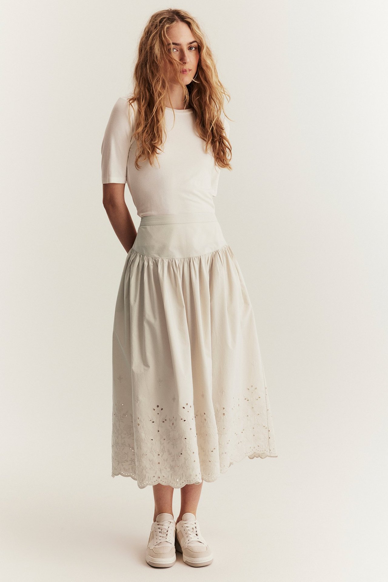 Vorderansicht: Frau trägt beigen Baumwoll-Midi-Rock mit Stickerei, weißes Kurzarm-Top und beige Sneaker.