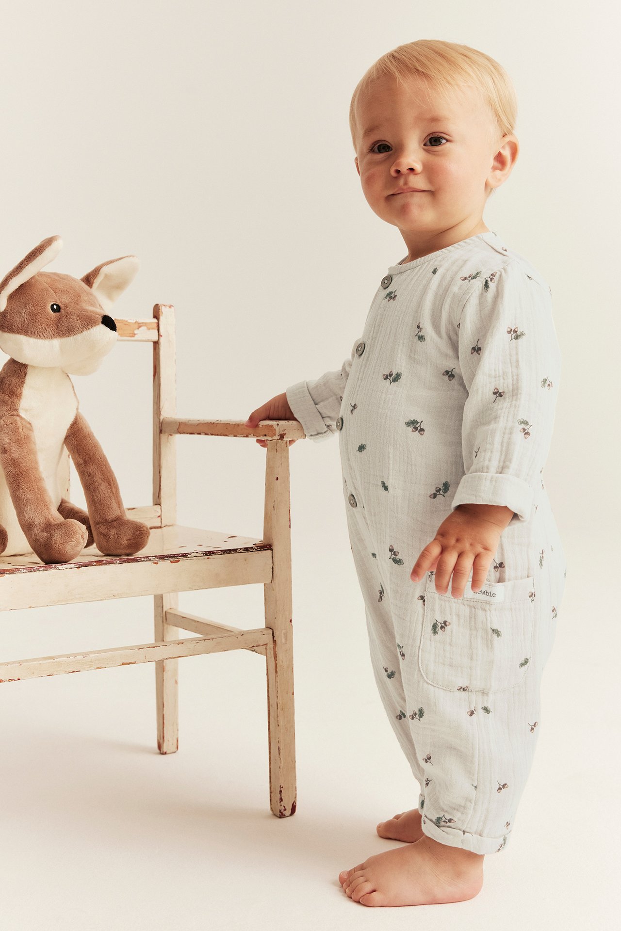 Seitenansicht eines Babys in beigem Baumwollmusselin-Playsuit mit Eichelmuster, hält Stuhl mit Fuchsspielzeug.