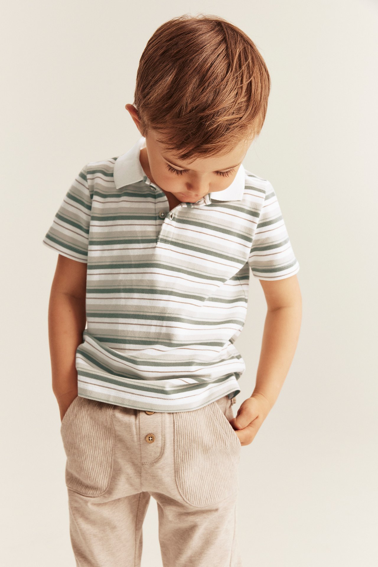 Vorderansicht eines Kindes in einem gestreiften Baumwoll-Poloshirt in Off-White, Grün, Braun und hellbeigen Hosen.