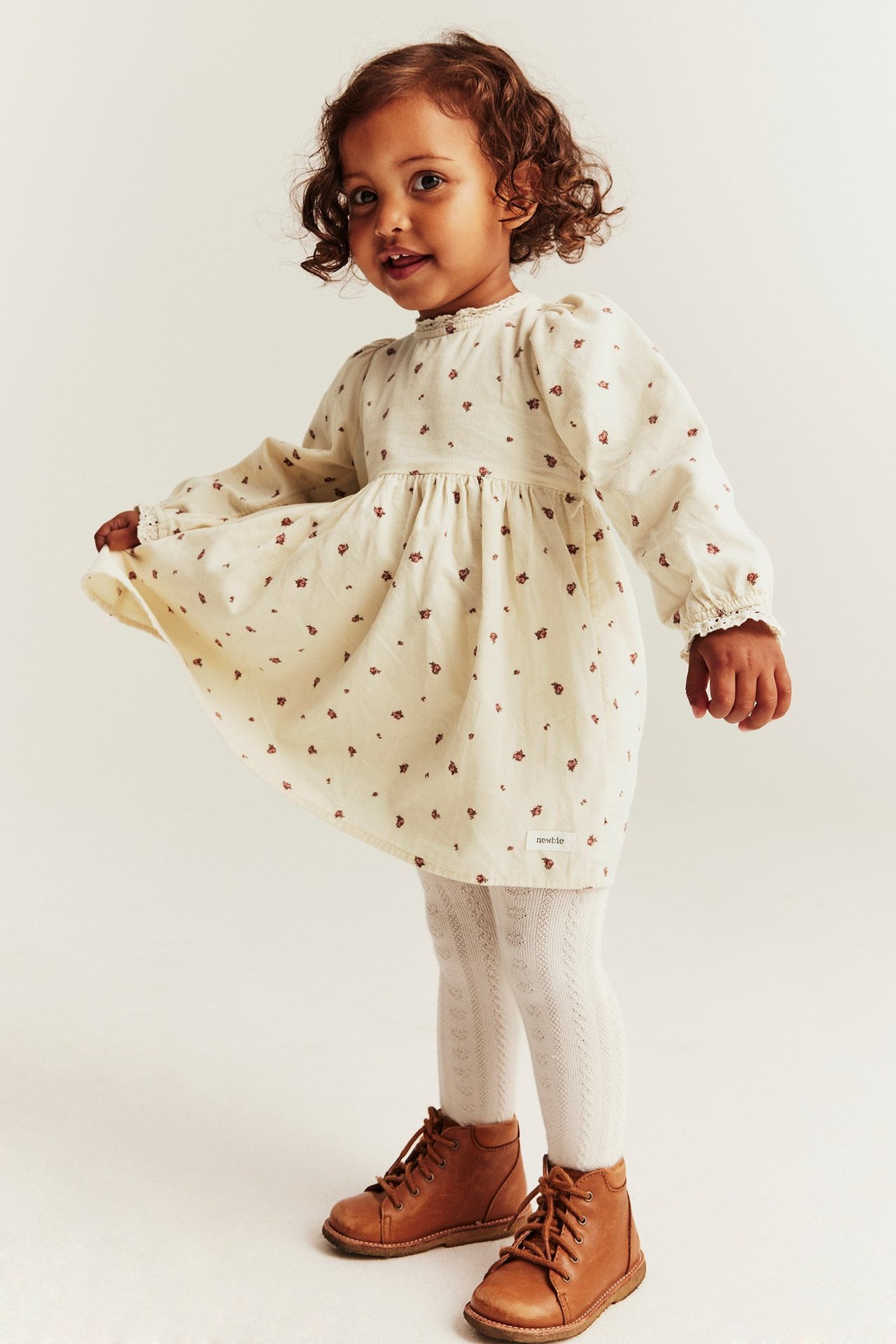 Kind trägt ein hellbeiges geblümtes Cordkleid mit Spitze, weiße gemusterte Strumpfhosen und braune Stiefel.