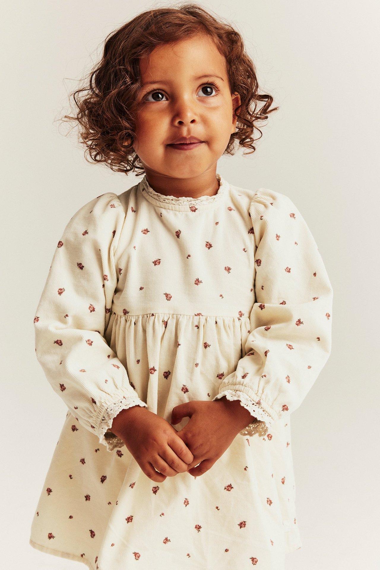 Vorderansicht: Kind in hellbeigem Blumen-Cordkleid mit Spitzenbesatz und Puffärmeln.