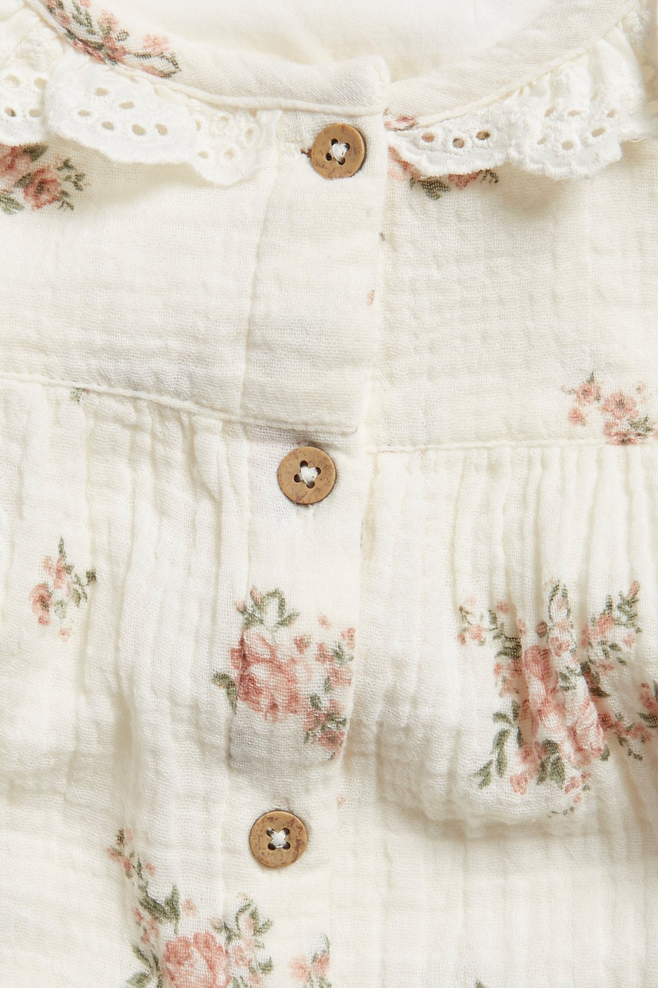 Nahaufnahme einer cremefarbenen Baumwollbluse mit Blumenmuster, Spitzenkragen und Holzknöpfen vorne.