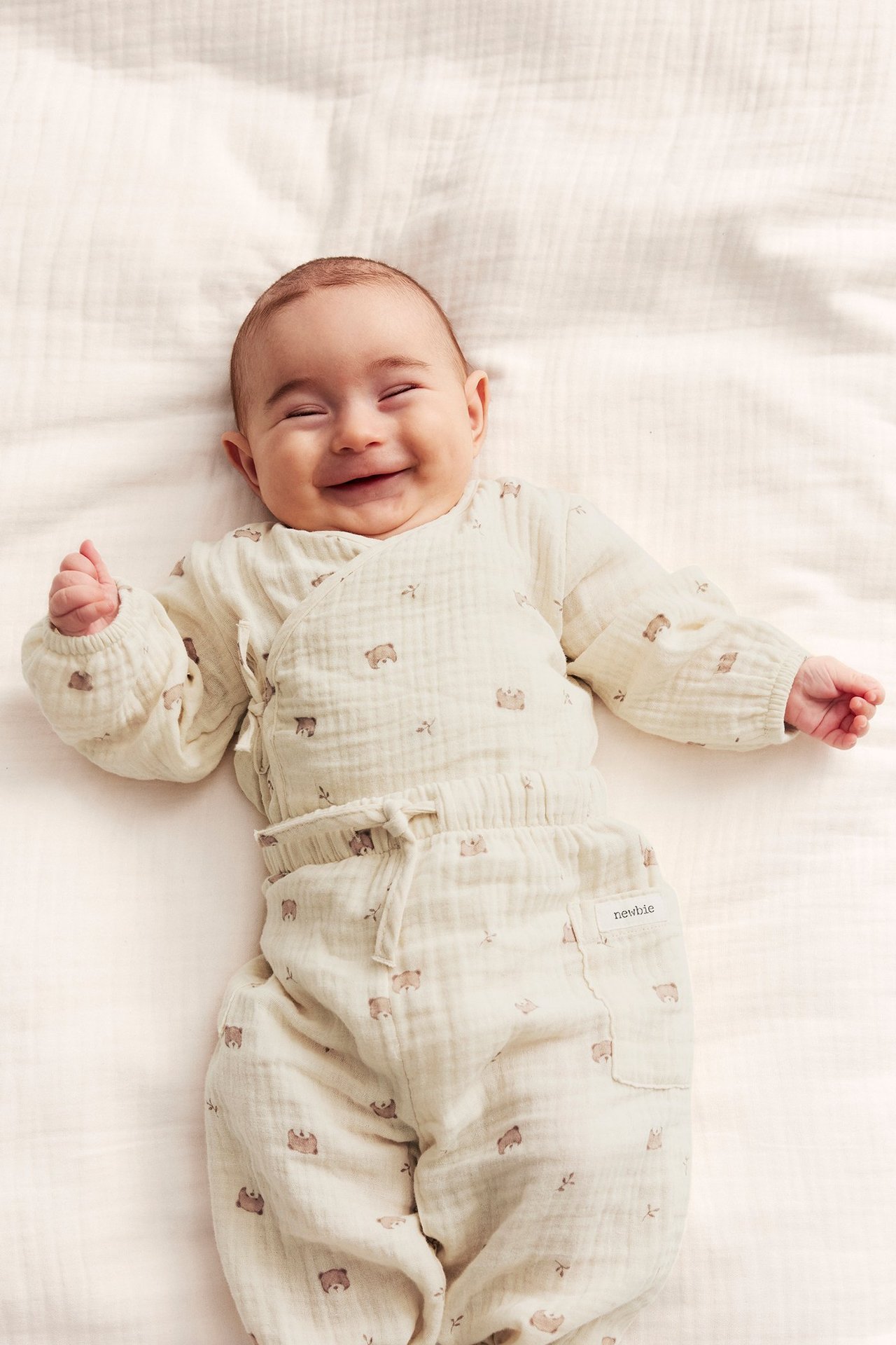 Top-down view of a smiling baby wearing a light beige muslin wrap bodysuit and matching pants with bear print.