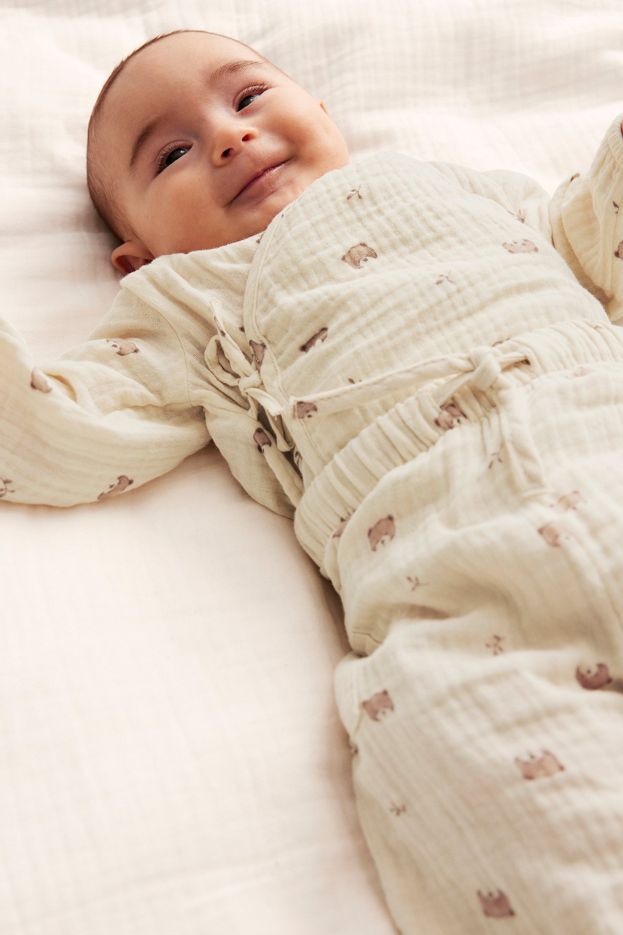 Front view: Smiling baby in light beige cotton muslin wrap bodysuit and matching bear-patterned pants.