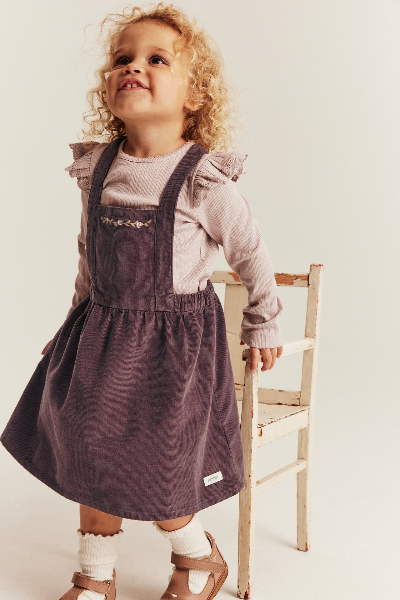 Kid's front view of a lilac corduroy pinafore dress with embroidery over a pink ribbed top.