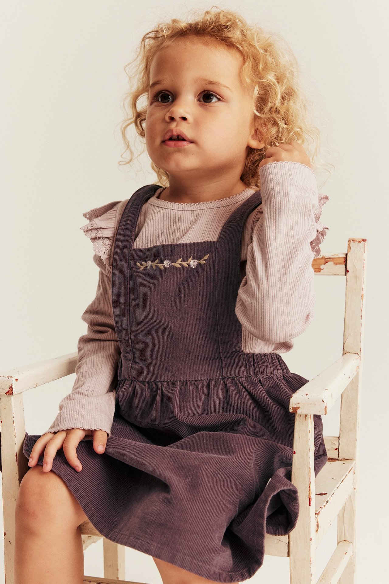 Front view of a kid wearing a lilac corduroy pinafore dress with floral embroidery over a light pink ribbed top.