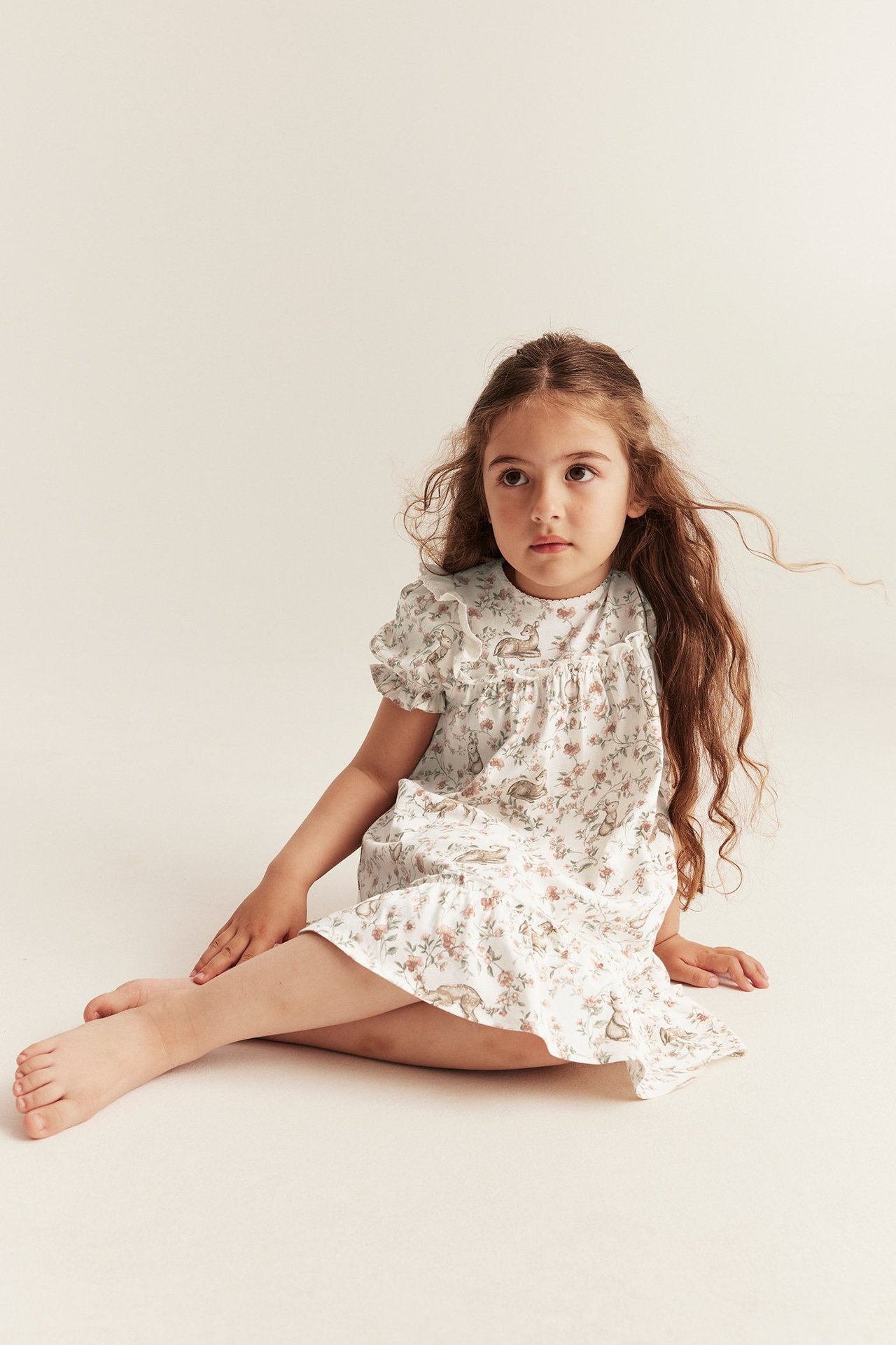 Front view of a kid wearing an off-white cotton jersey dress with a rabbit and floral pattern, puff sleeves, and ruffles.