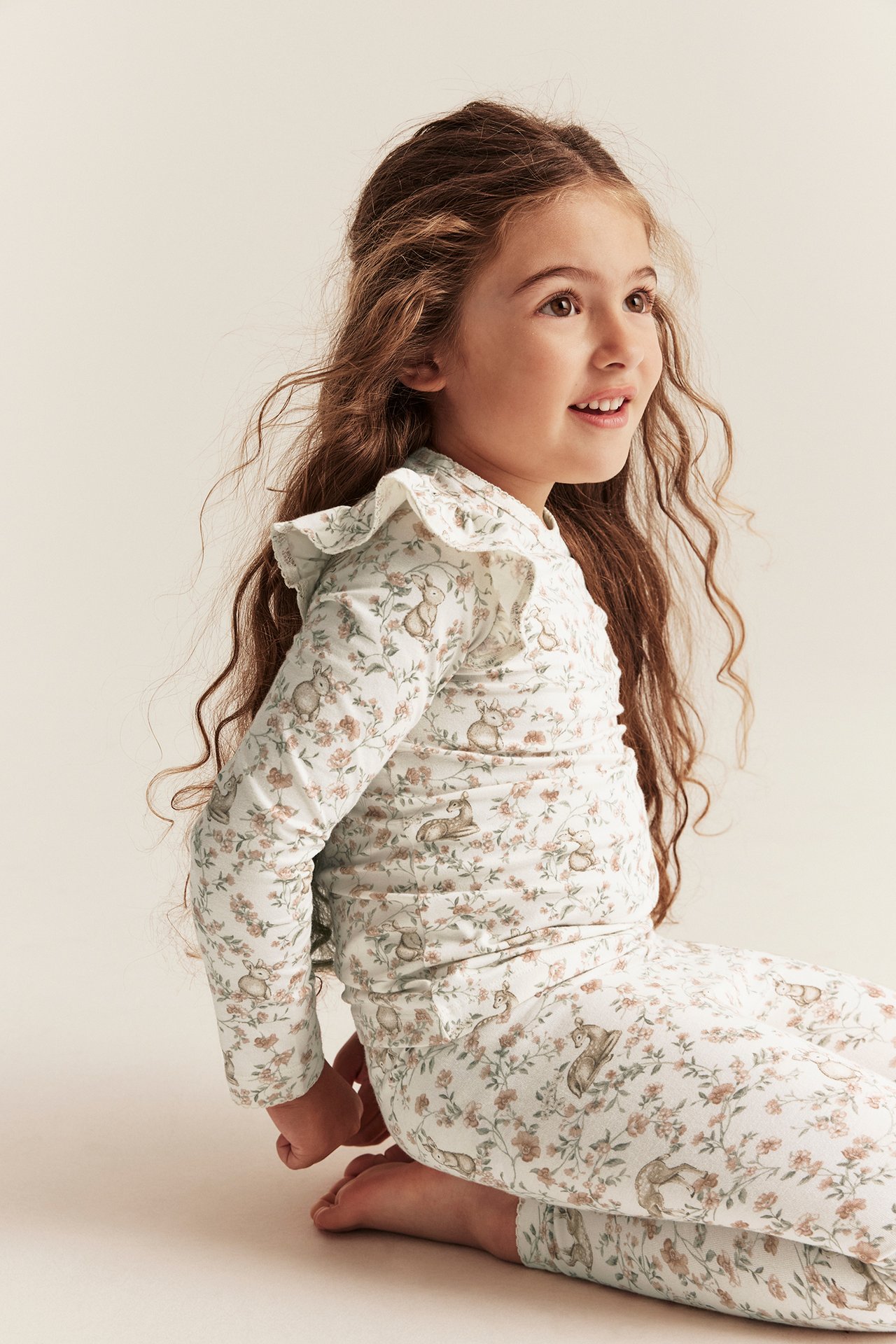 Side view: Kid girl in off-white floral animal print long-sleeved top with ruffles, matching pants.