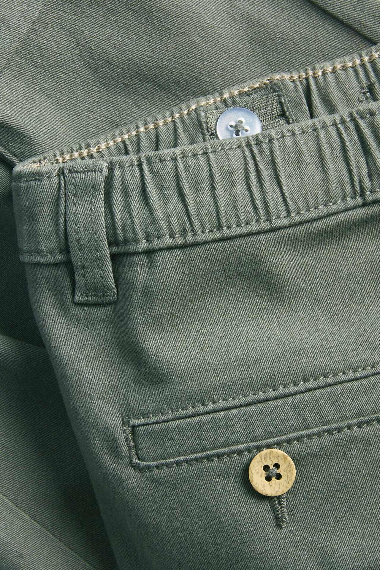 Close-up of dark green woven pants, showing elastic waist with adjustable button and decorative back pocket.