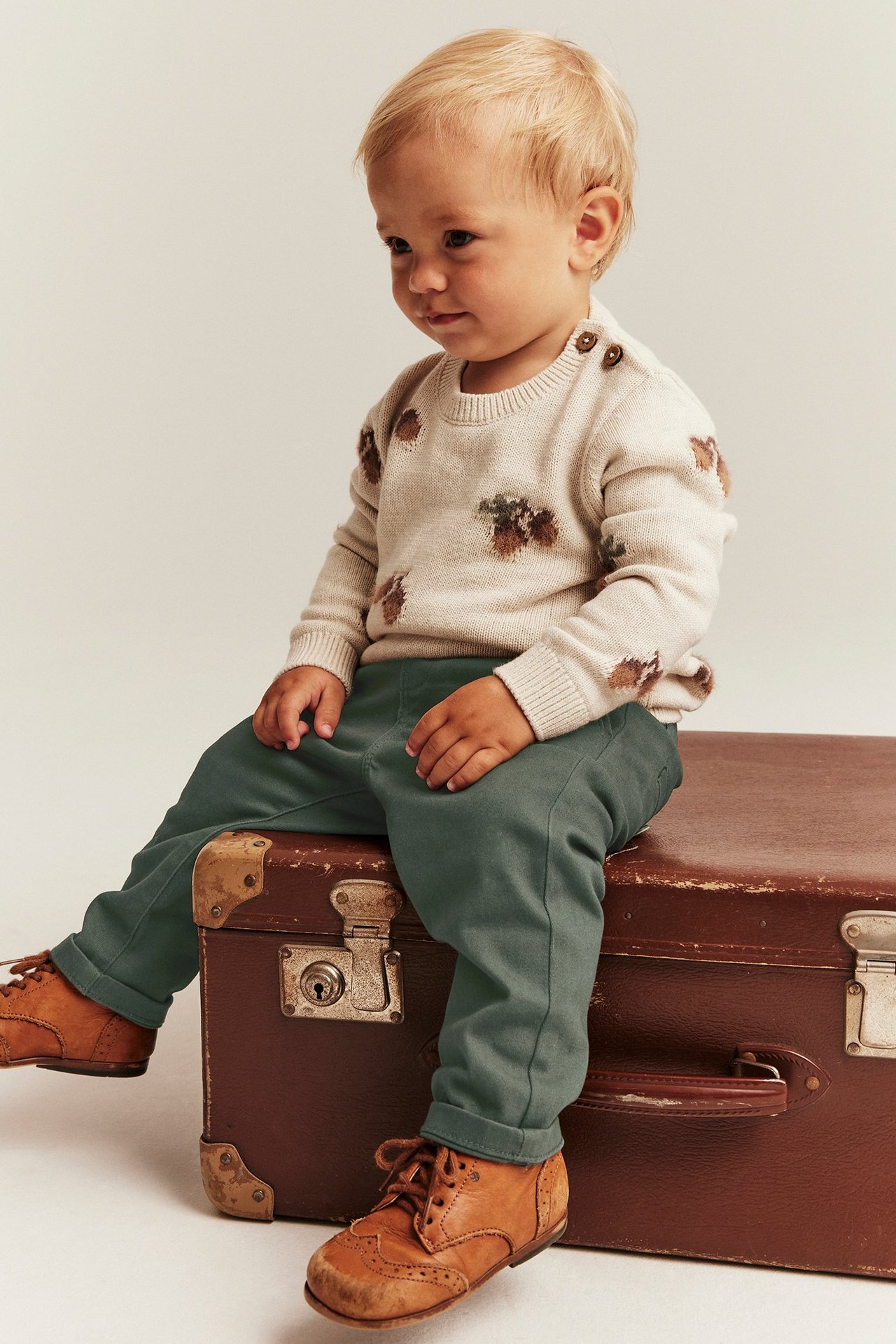Front view of a baby wearing dark green woven trousers, a beige patterned sweater, and brown lace-up shoes.