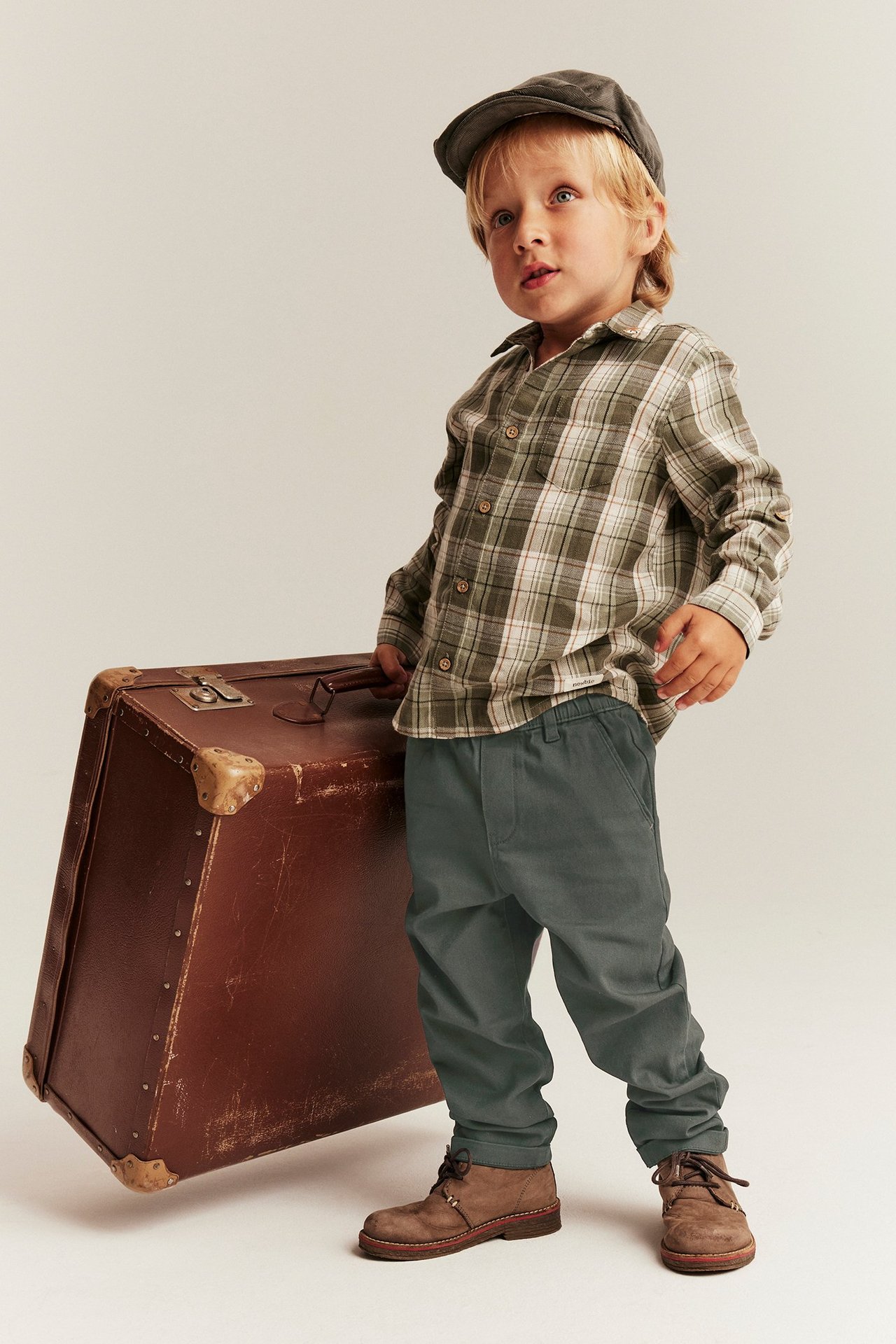 Vorderansicht eines Kindes in dunkelgrünen Webhosen, einem karierten Hemd, einer dunklen Mütze und braunen Schuhen.