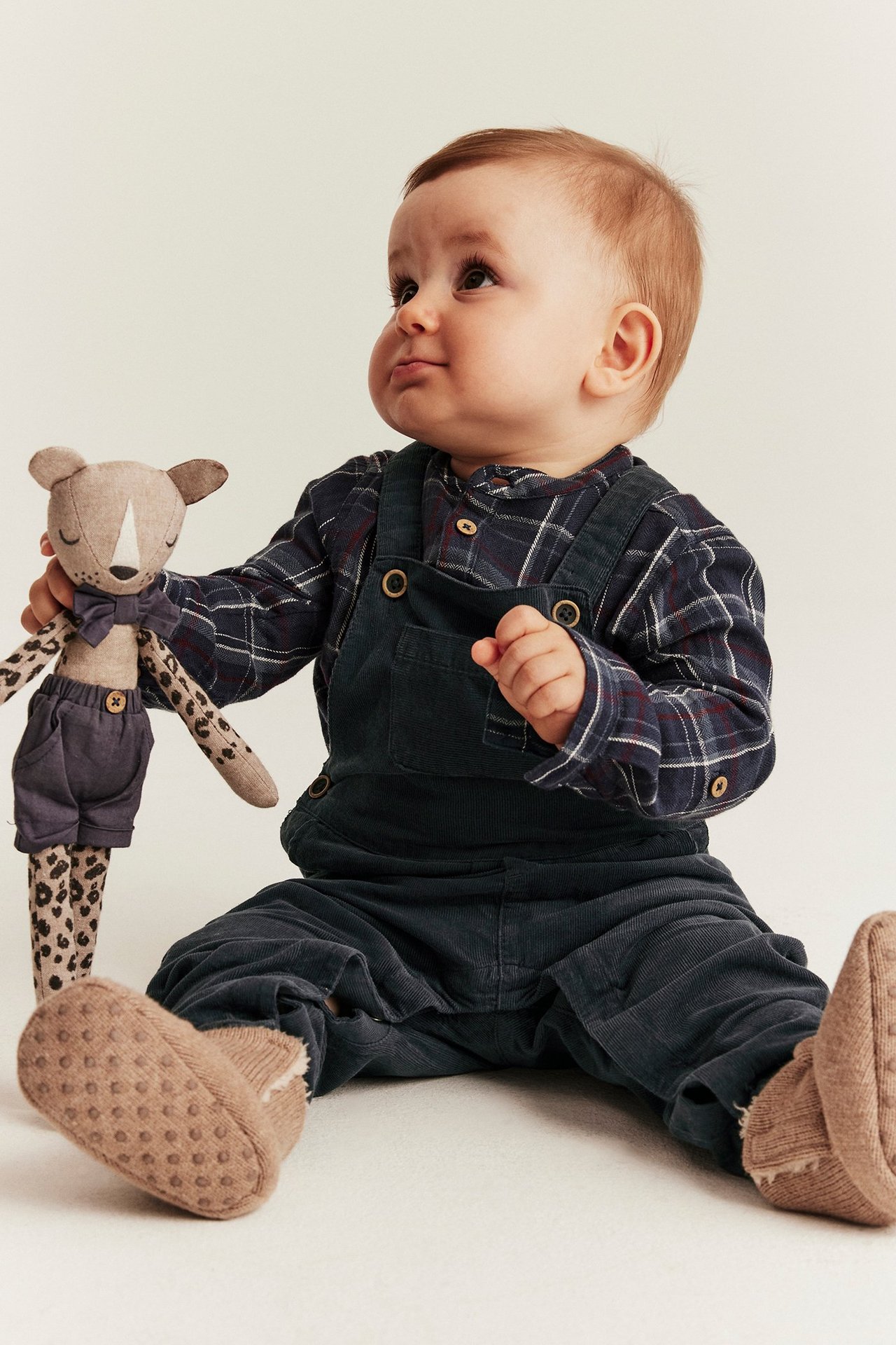 Baby in dark blue dungarees in cotton, sitting with a toy.