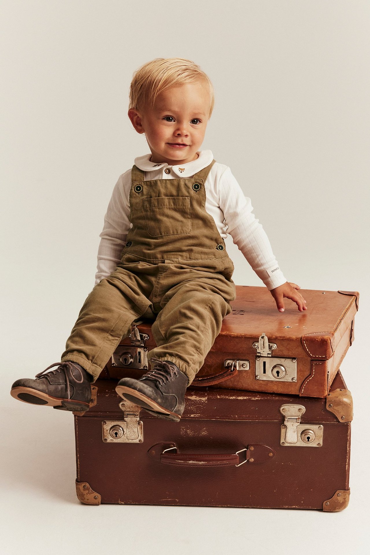 Kid in brown Manchester overalls with adjustable straps, sitting on vintage suitcases, front view.